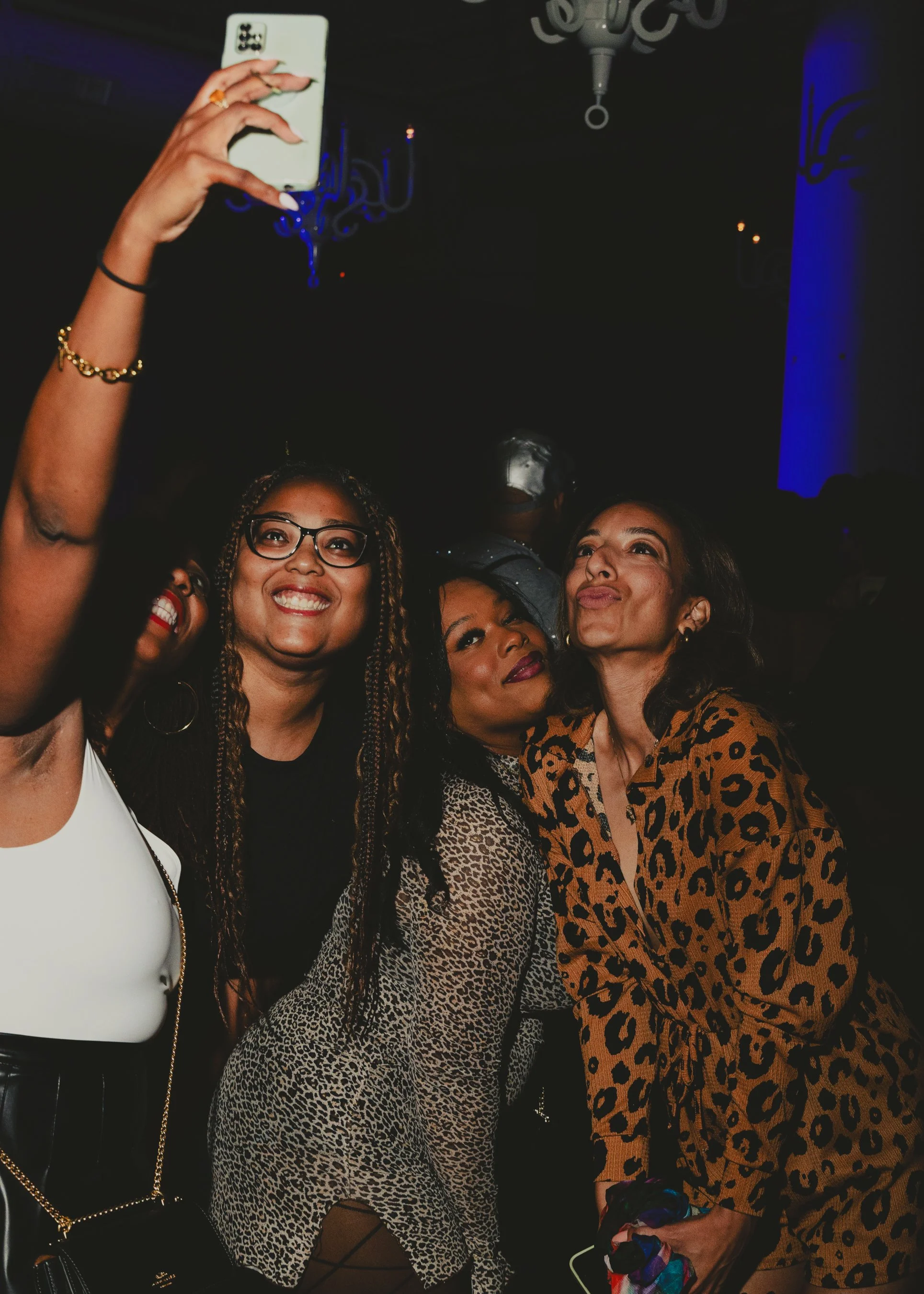 Group of women taking a selfie at a party or event, with dark background and blue lighting, some smiling and posing in fashionable clothing.
