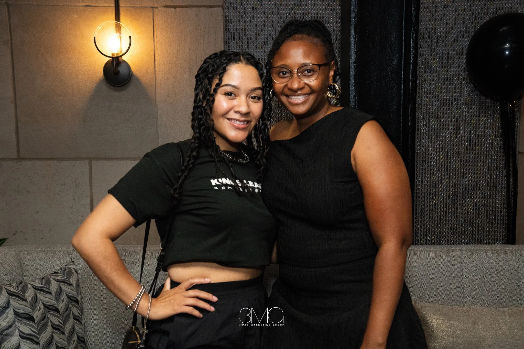 Two smiling women standing close together in a room, one with braided hair and glasses, wearing a black top, and the other with curly hair, wearing a black t-shirt and earrings.
