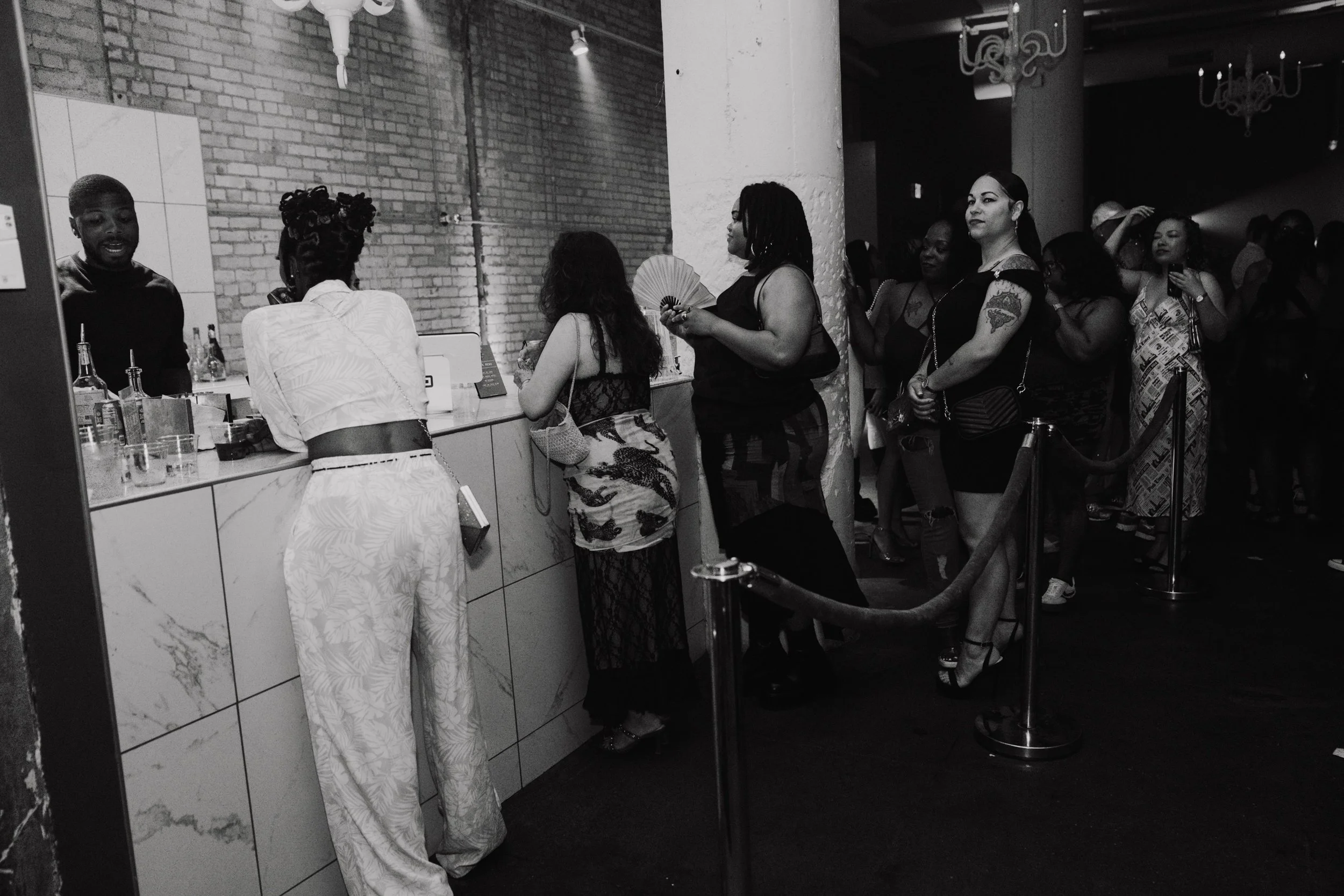 A group of women standing in line at a bar or drink counter inside a dimly lit venue with exposed brick walls. Some women are holding drinks, one has a fan, and the scene suggests an event or social gathering.