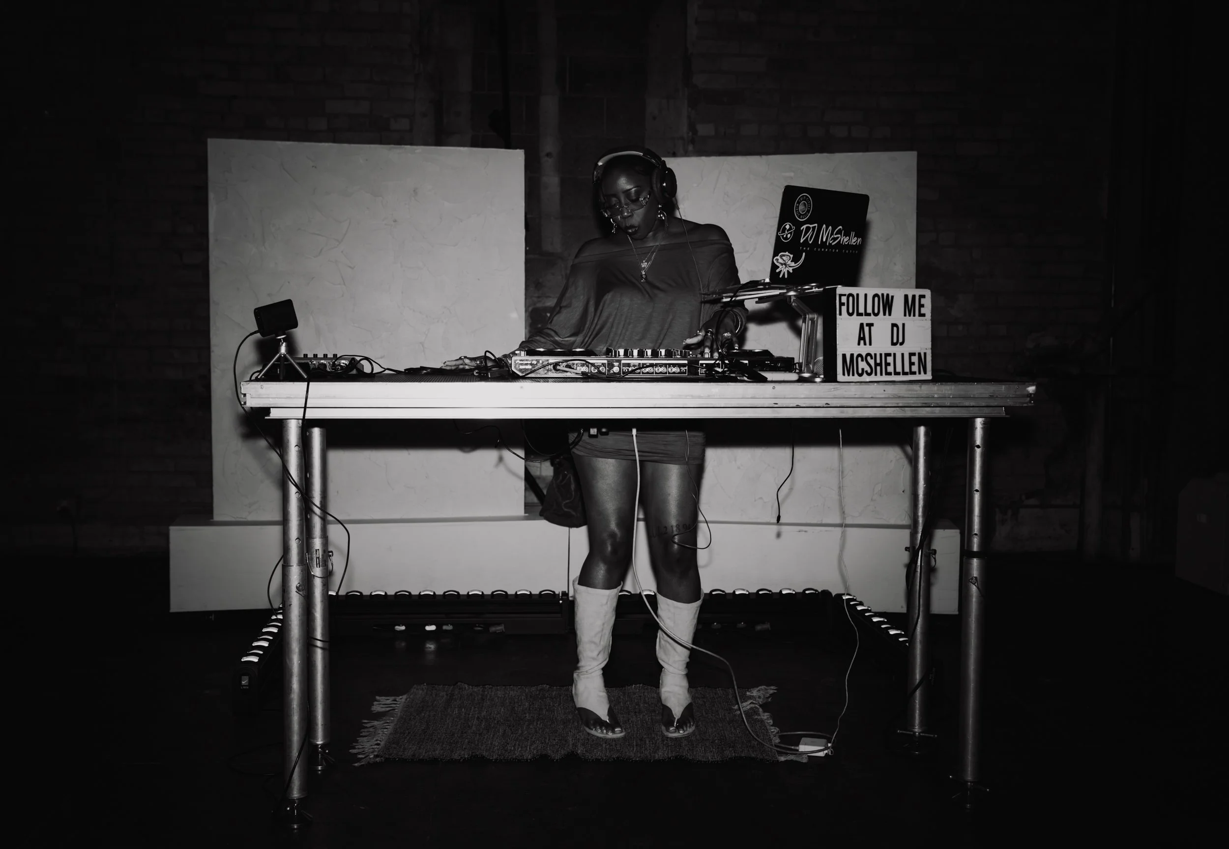 A woman DJ wearing headphones, glasses, patterned top, and boots, standing behind DJ equipment with a sign that reads "Follow me at DJ MCSHellen."
