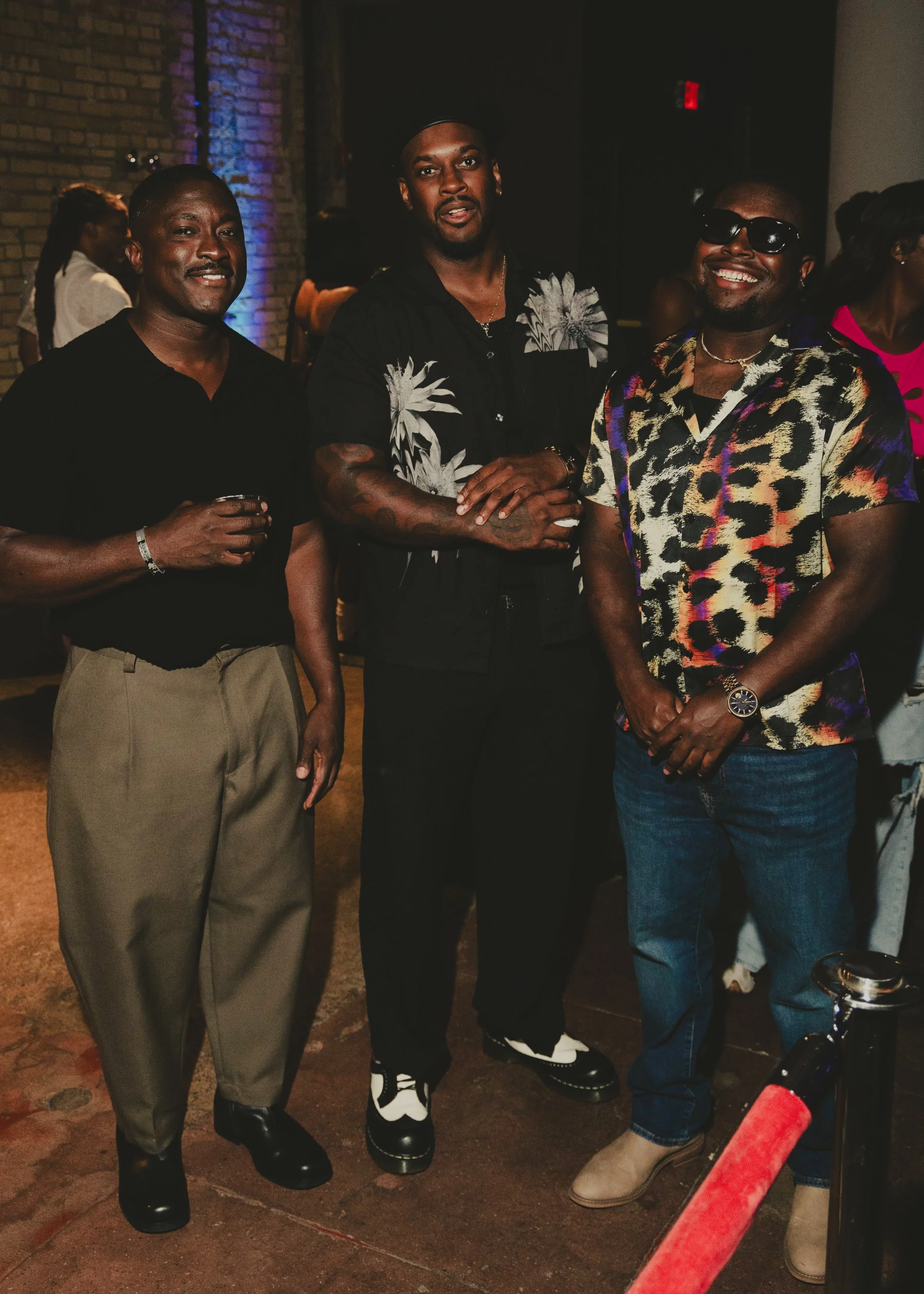 Three men standing together at a social event, dressed casually, with a brick wall and other people in the background.