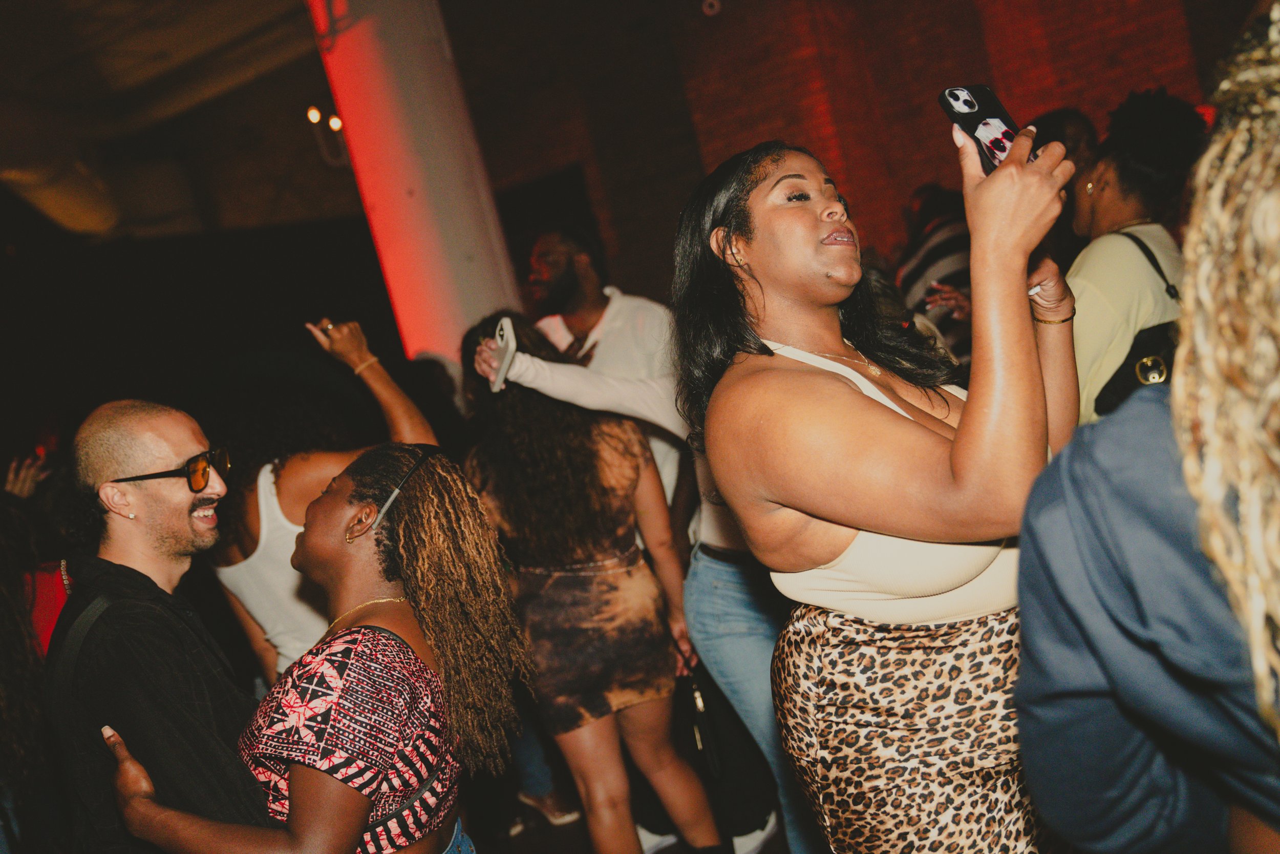 Group of people at a lively indoor event, some dancing and taking photos, with diverse skin tones and stylish clothing.