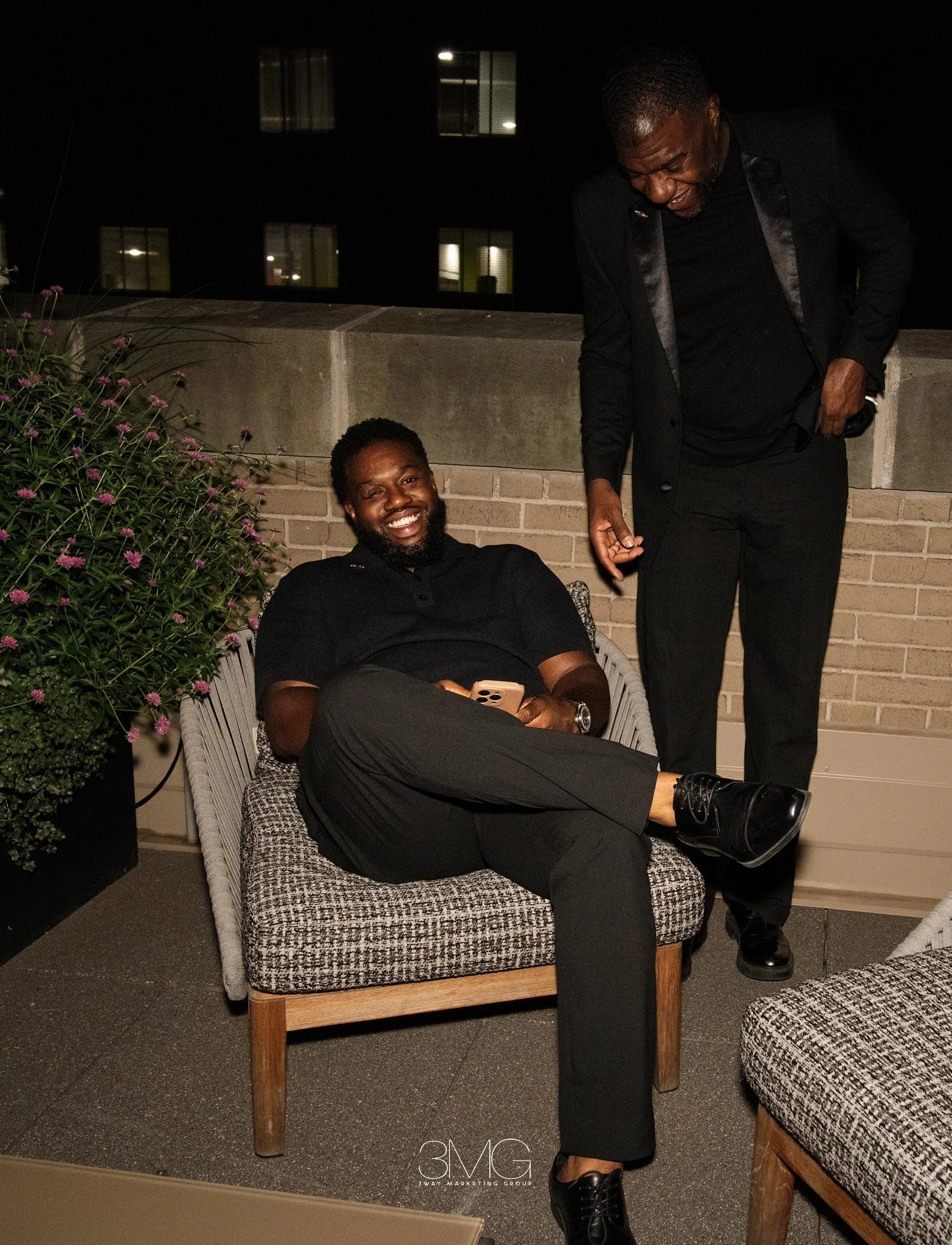 Two men in black suits are on a rooftop patio at night, with one sitting on a bench smiling and holding a phone, and the other standing and laughing, nearby plants, and buildings with lit windows in the background.