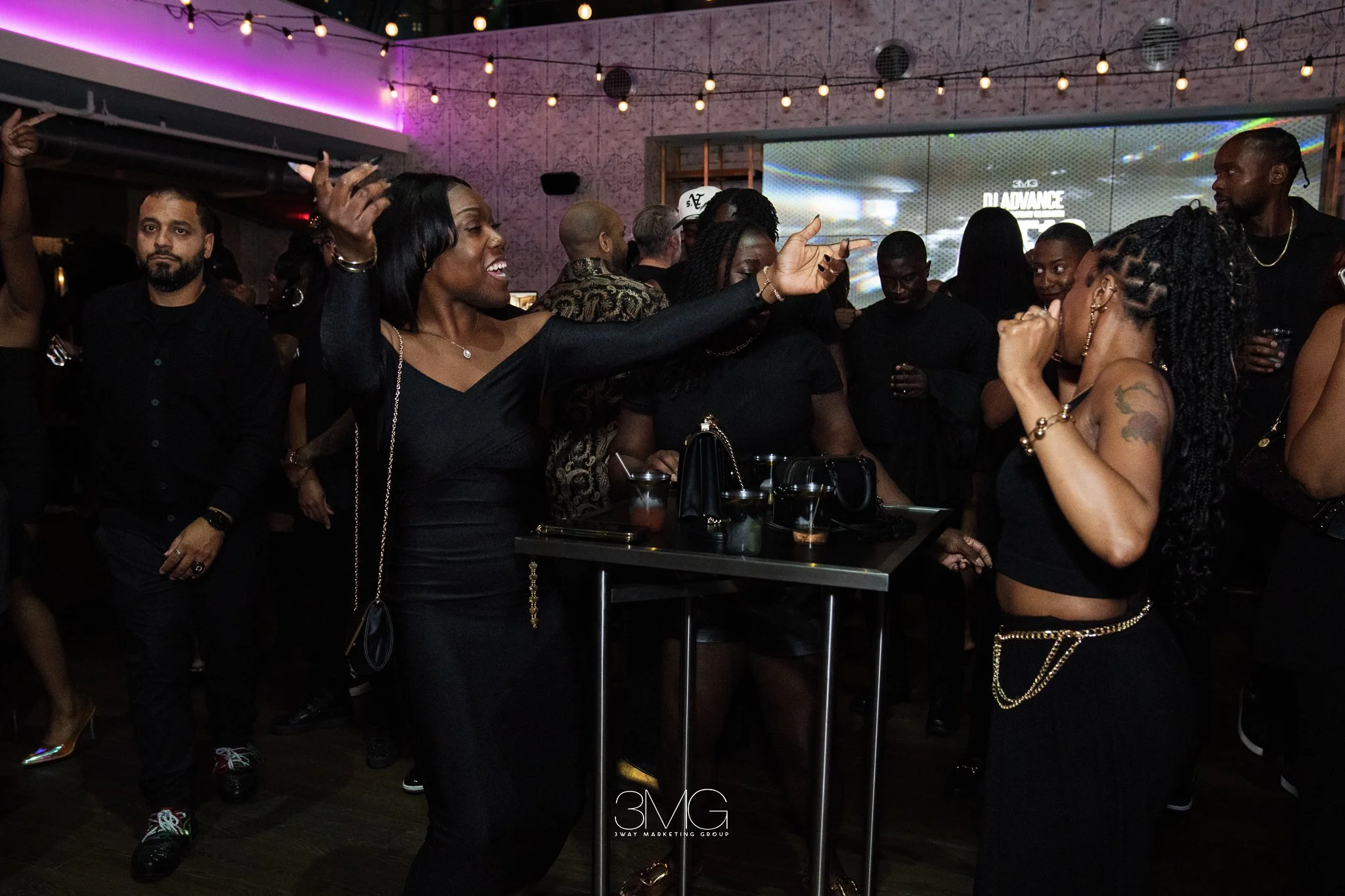 A group of people dancing and socializing at a party or nightclub with colorful lighting and a screen in the background displaying the logo '3MG'.