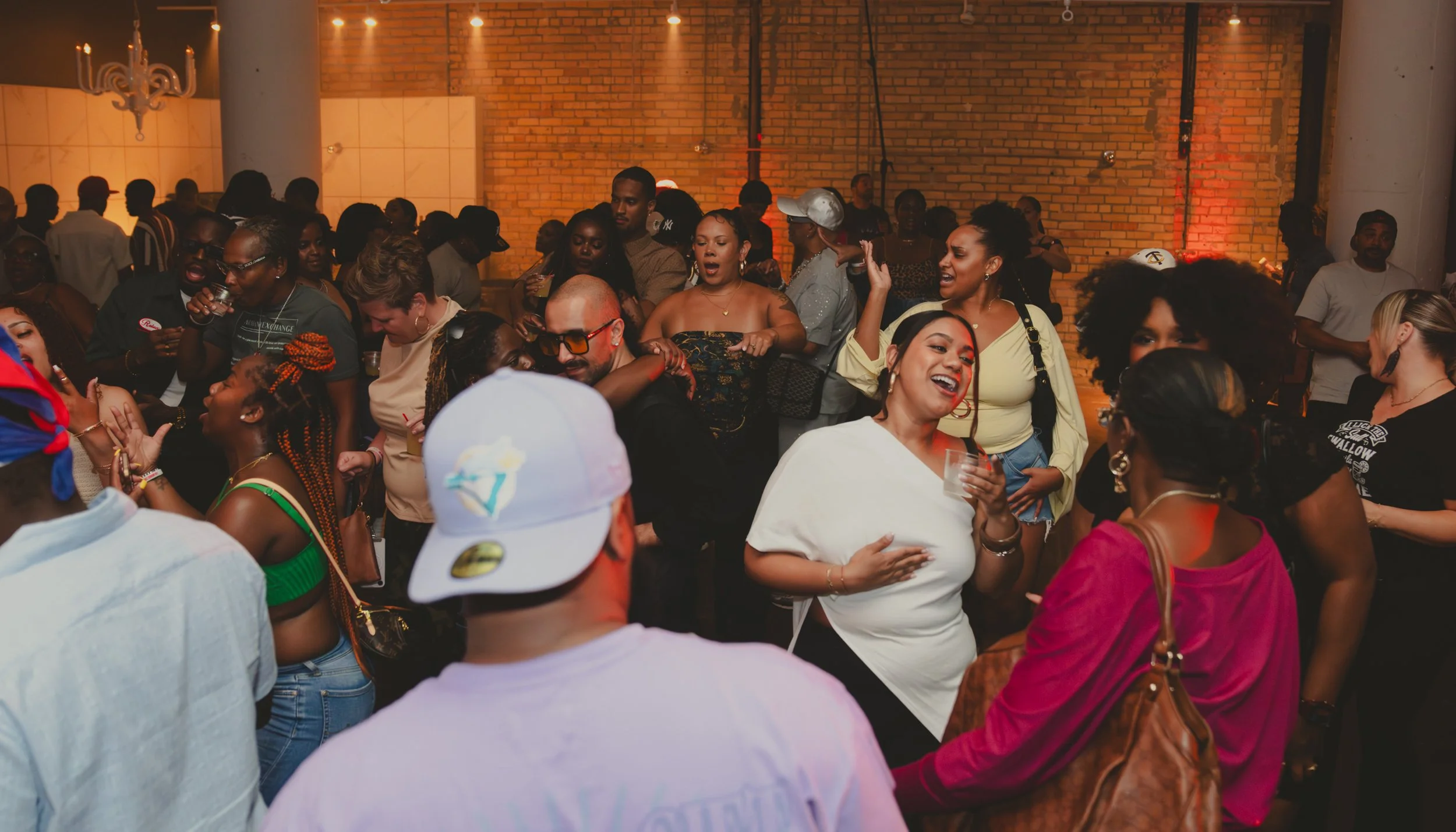 A lively indoor party with many people dancing and socializing against a brick wall, with warm lighting and some people holding drinks.