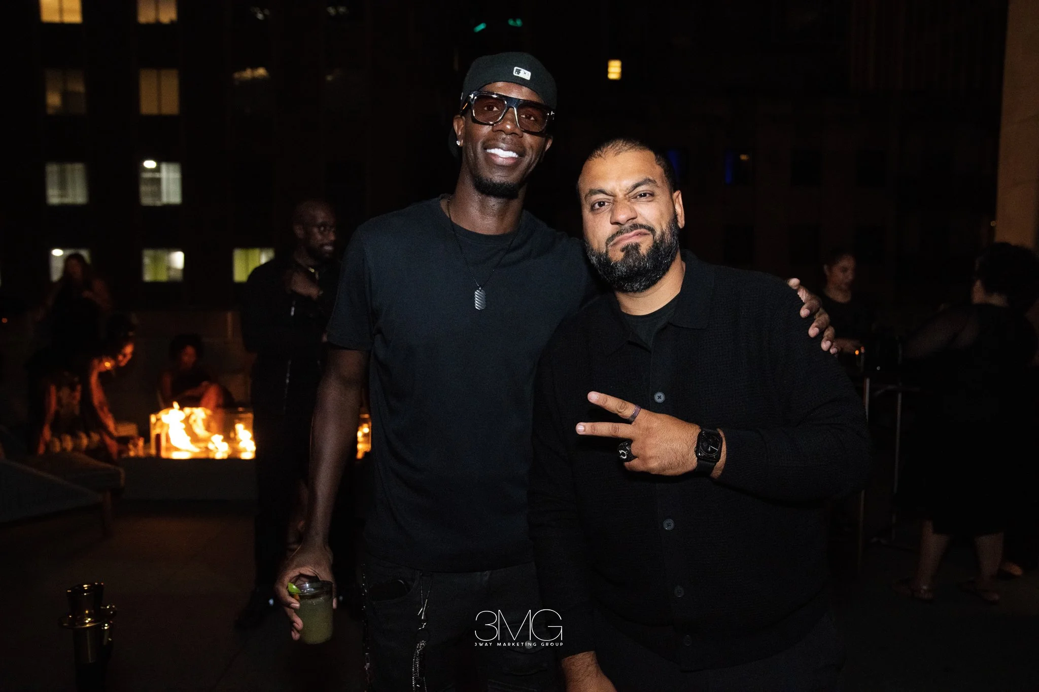 Two men posing together at night, one wearing a black cap and glasses, the other with a beard and black shirt, in an outdoor setting with people and lights in the background.