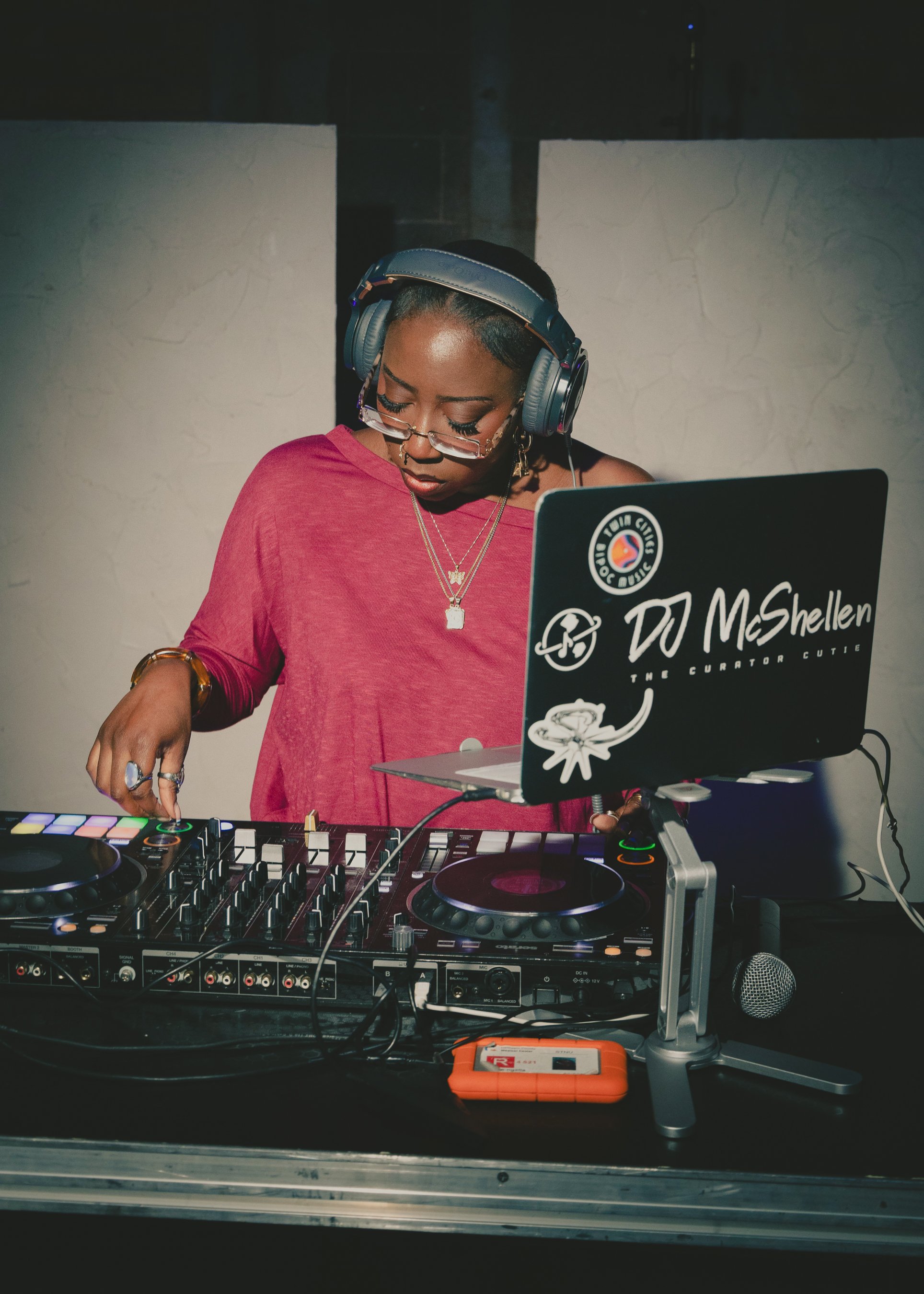 A female DJ wearing glasses and headphones, working with a DJ mixer and laptop on a table.