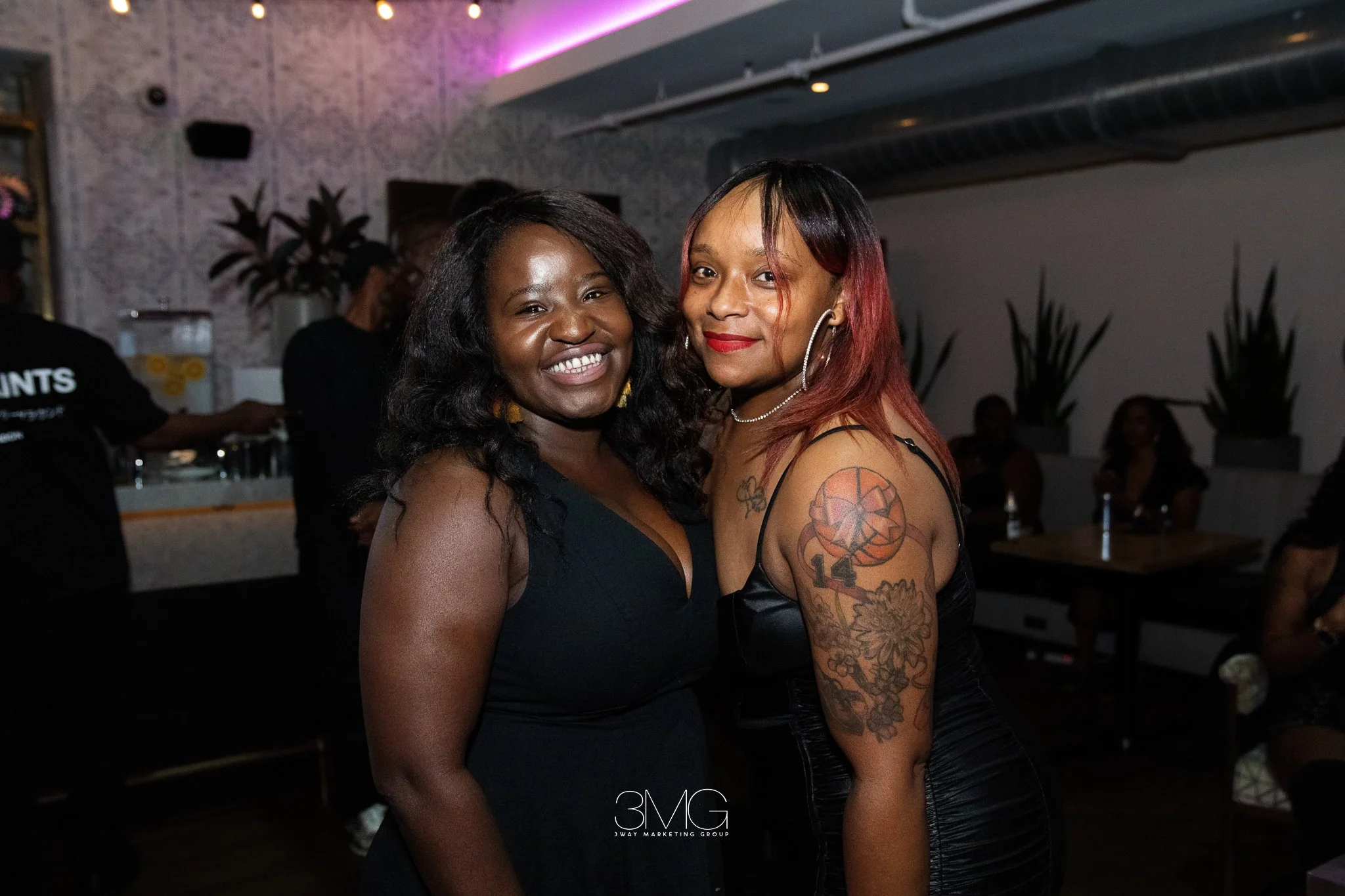 Two women smiling at a social event, standing close together in a dimly lit indoor venue with people and potted plants in the background.