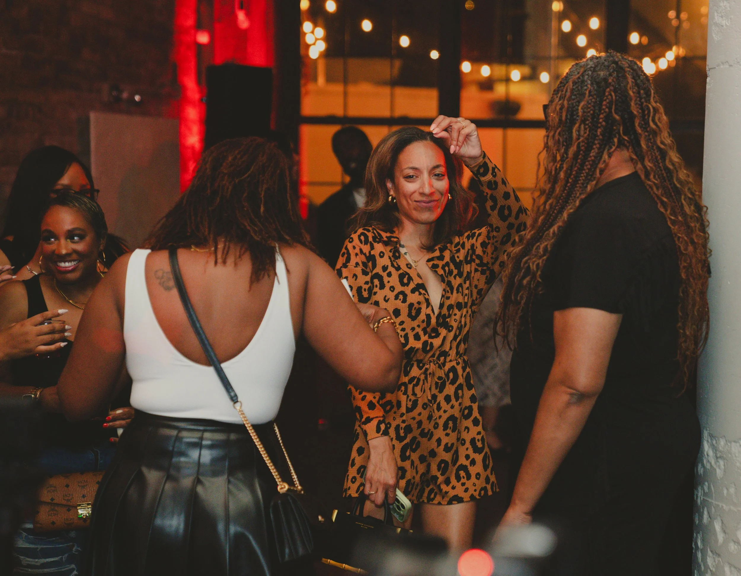 A group of women at a social gathering indoors, with warm lighting and brick walls, dressed stylishly and engaging in conversation.