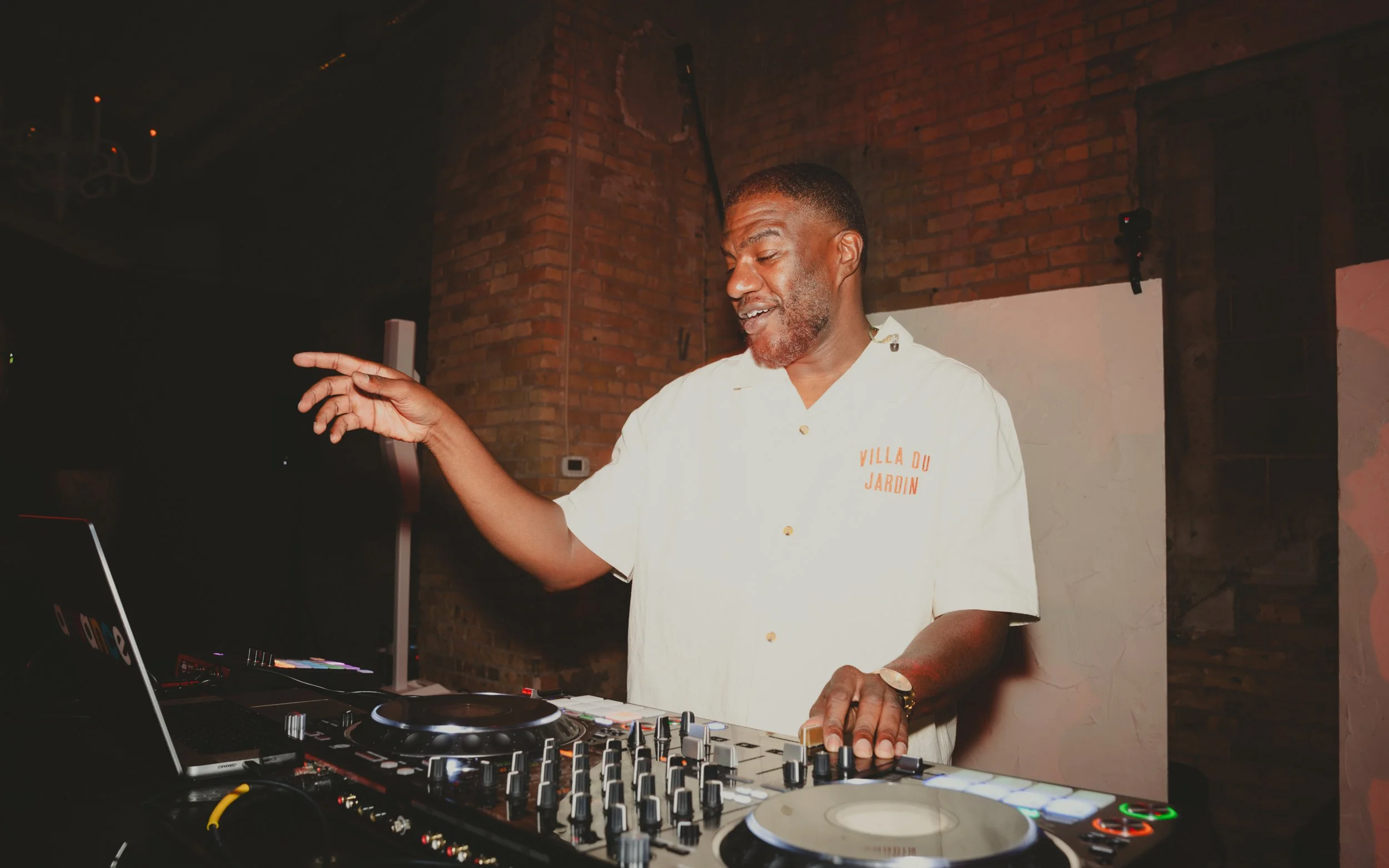 A man with gray hair and beard wearing a cream-colored shirt, DJing with turntables and a mixer in a dimly lit room with brick walls.