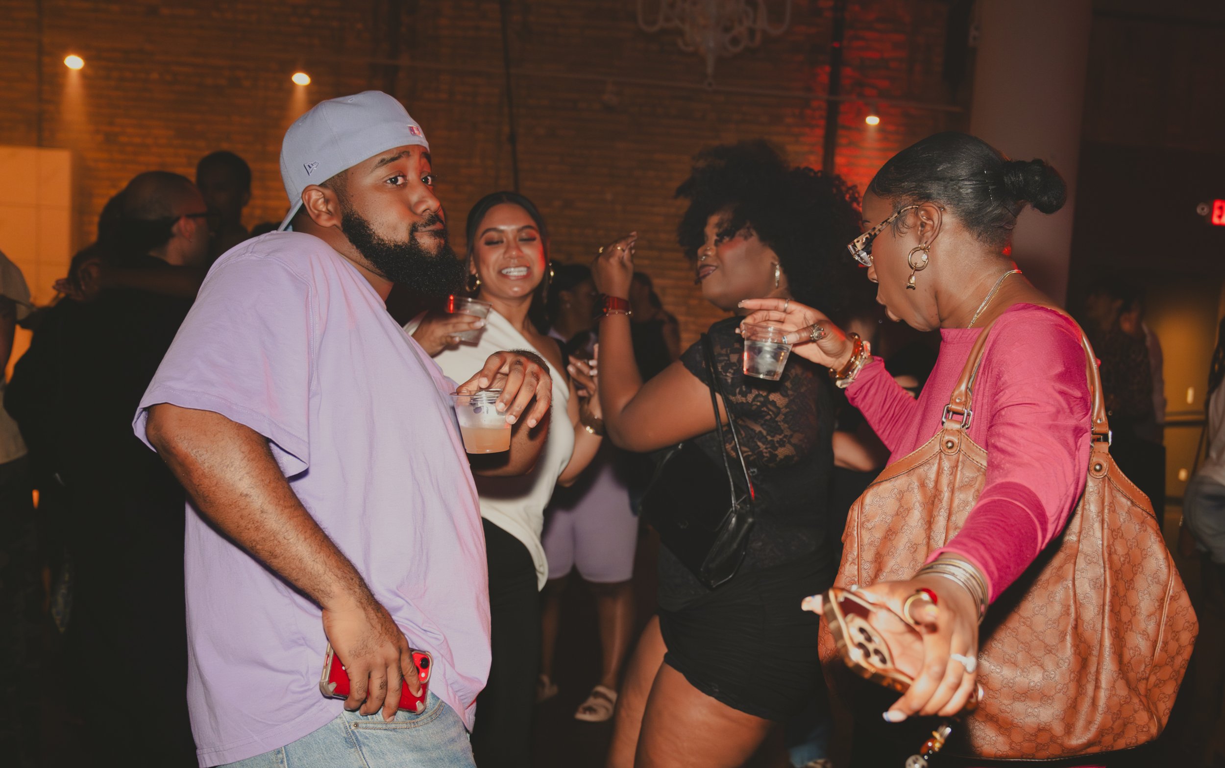 Group of people socializing at a bar or club, holding drinks, with warm lighting and brick wall background.