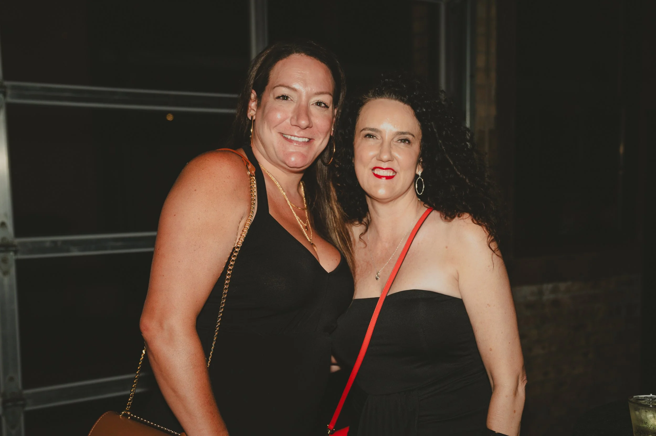 Two women smiling at a nighttime event, dressed in black dresses, with one wearing gold jewelry and the other with large hoop earrings and red lipstick.