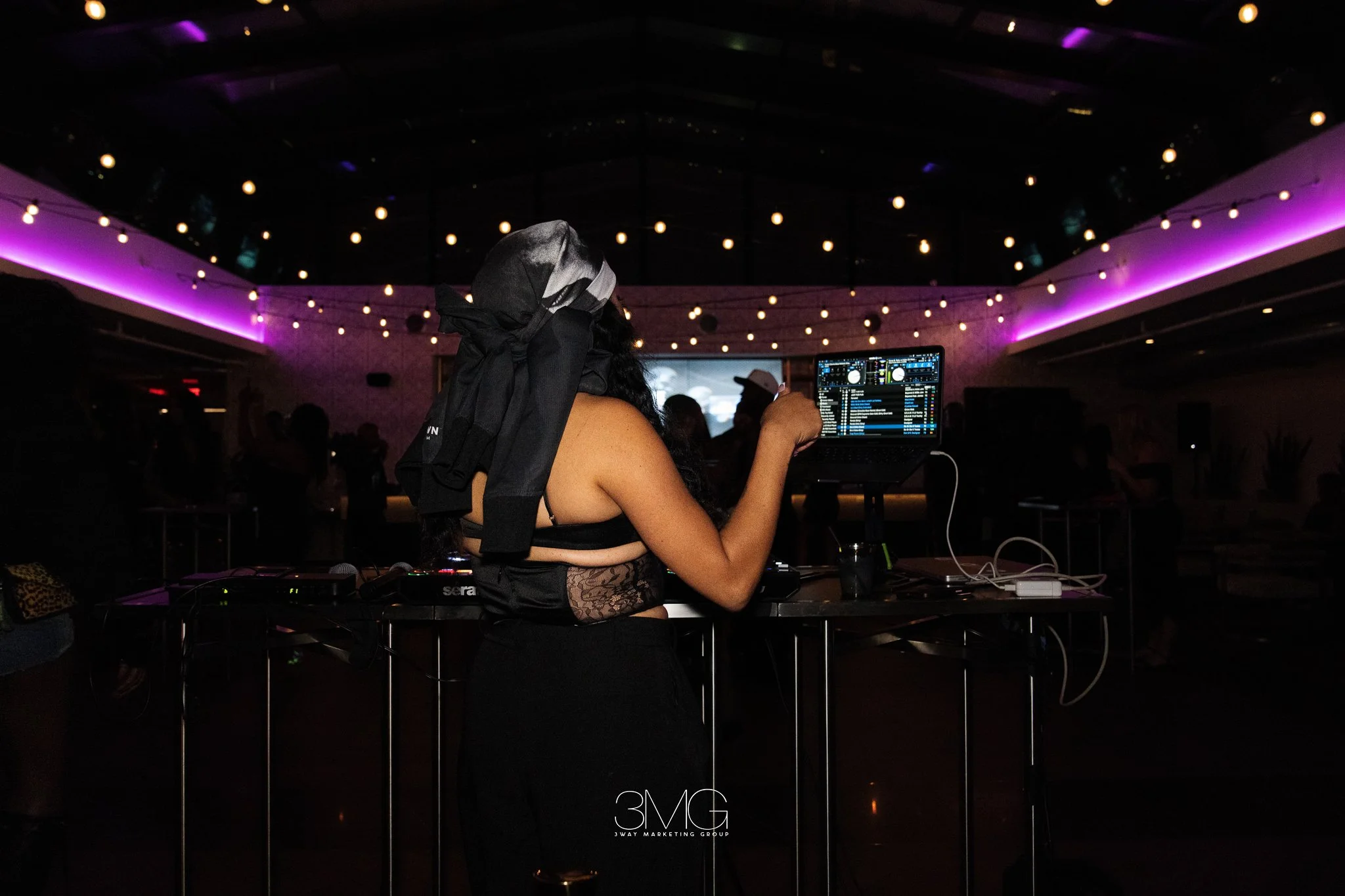 A woman DJ wearing a black headscarf and lace top mixing music at an event with purple lighting and string lights overhead, with the logo '3MG' at the bottom center.