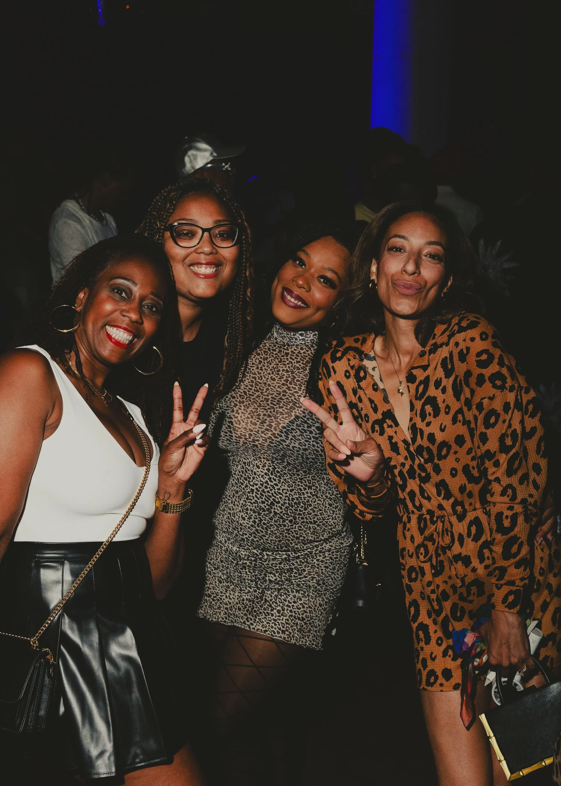 Four women smiling and posing for a photo at a social event, with dark background and subtle blue lighting.