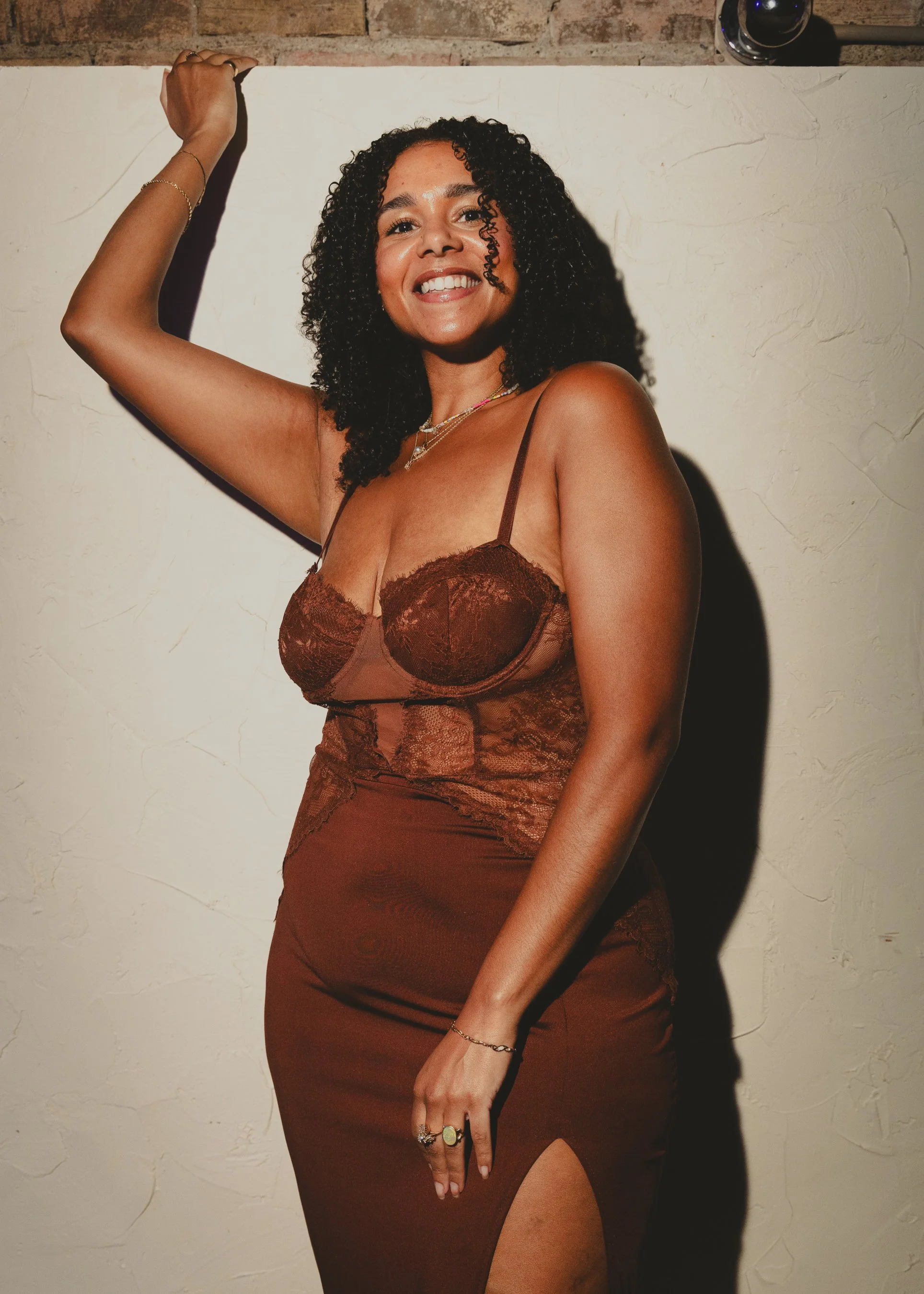 A woman with curly black hair smiling, wearing a brown lace dress with spaghetti straps, jewelry, and a ring, standing against a cream textured wall with a brick upper section.