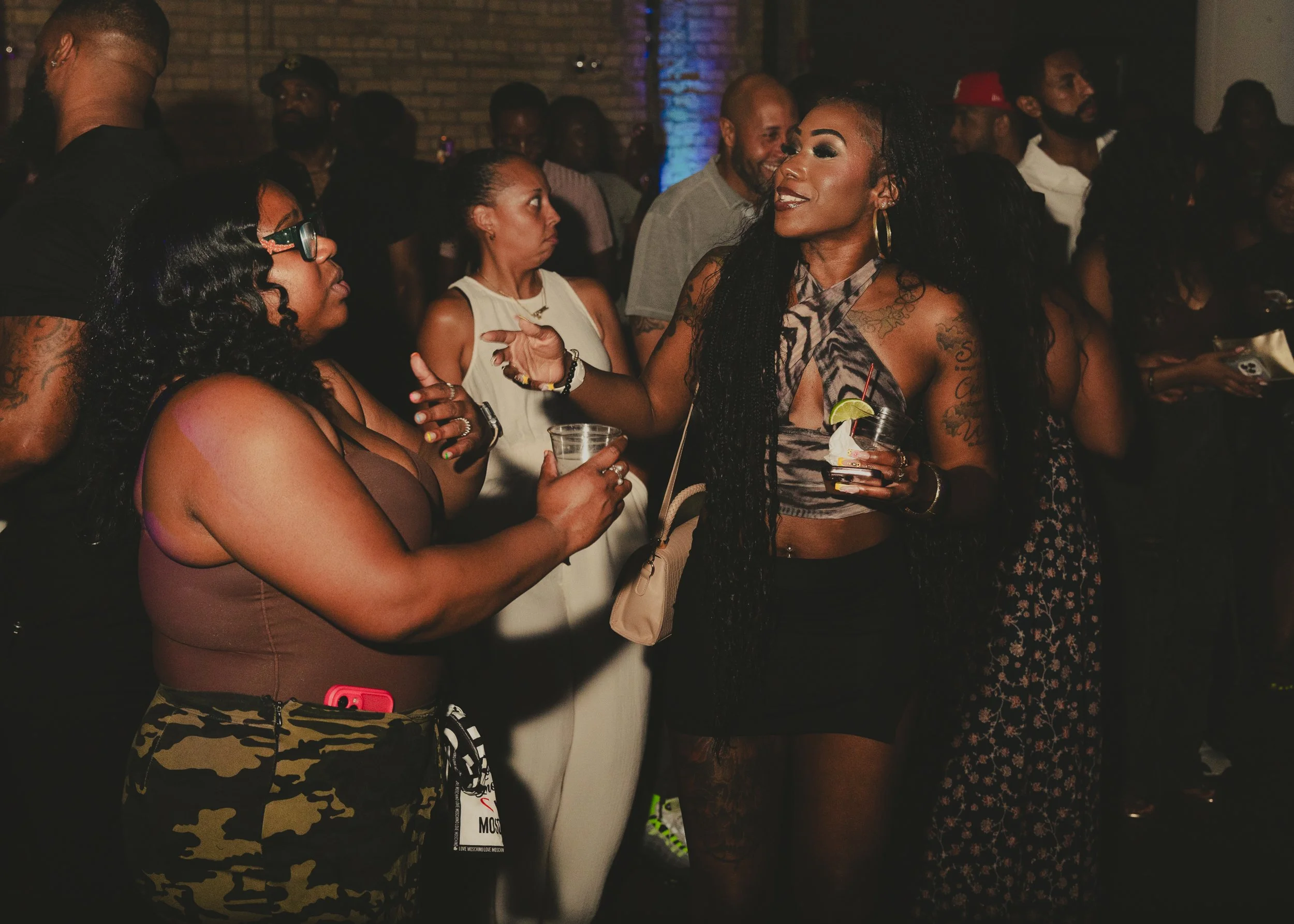 Group of people socializing at a party in a dimly lit venue, some holding drinks, with brick walls in the background.