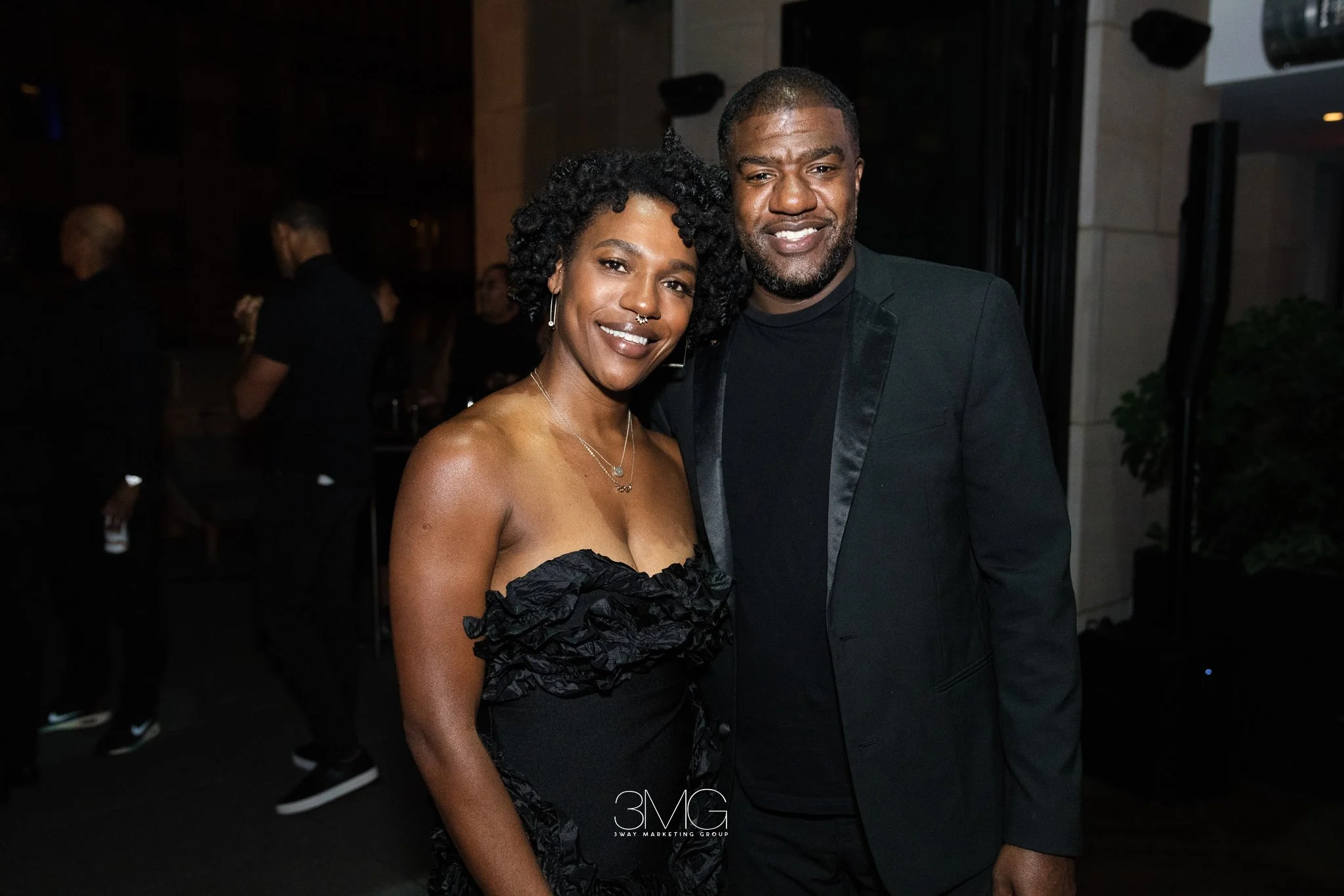 A man and woman smiling at a party or event, standing close together. The woman is wearing a strapless black dress with ruffled details, has curly hair, and is wearing jewelry including earrings and a nose ring. The man is wearing a black suit jacket