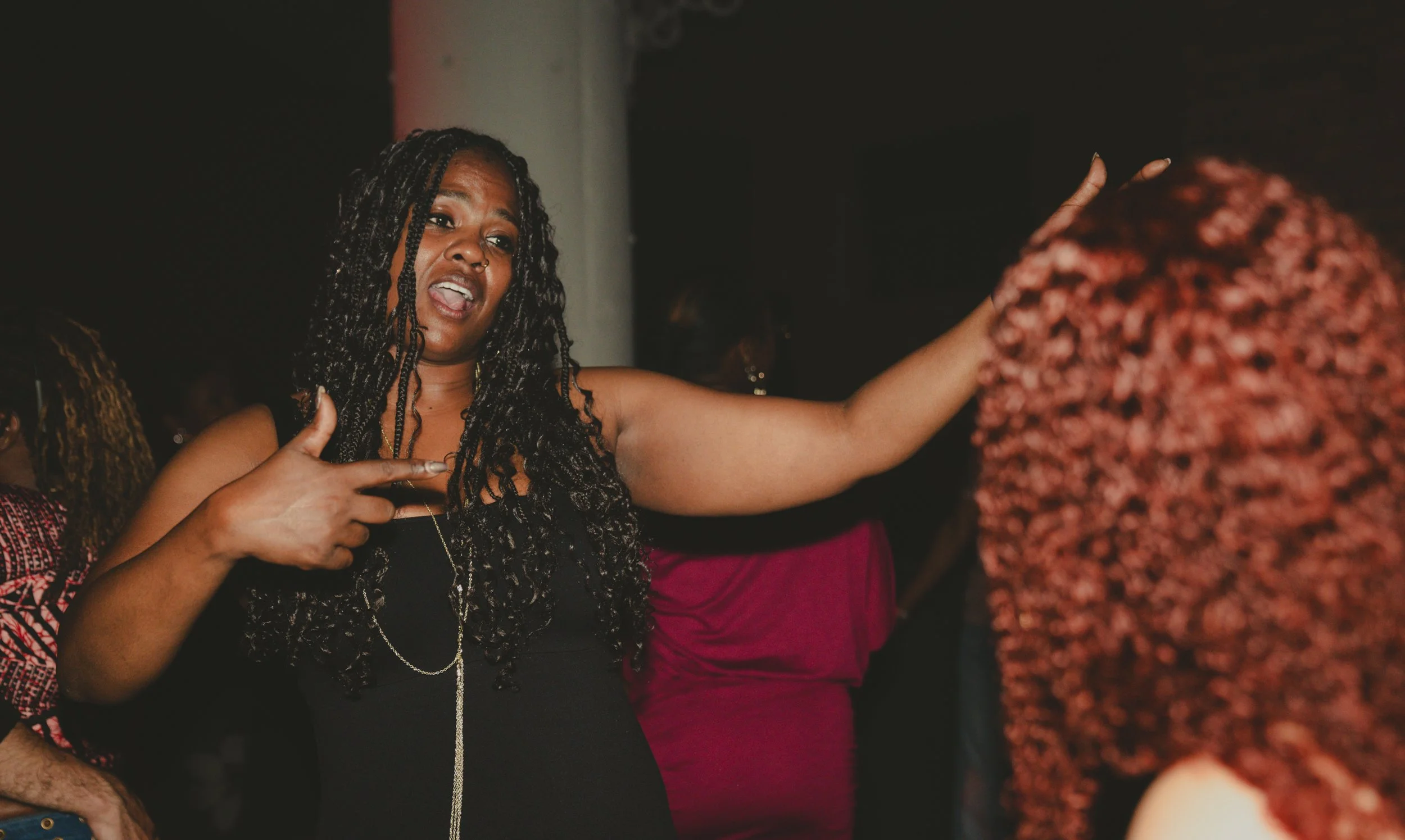 A woman with long curly hair, pointing at herself and speaking, at a social gathering, with other people around.