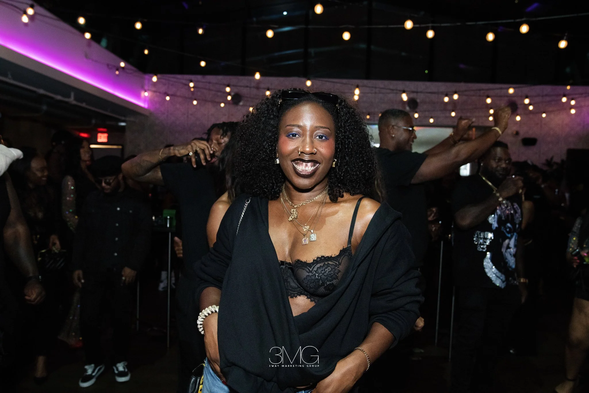 A woman with curly black hair and dark lipstick smiling at a lively indoor party. She wears a black lace top, layered necklaces, a white bracelet, and sunglasses on her head. The background shows other party guests dancing and enjoying the music, wit