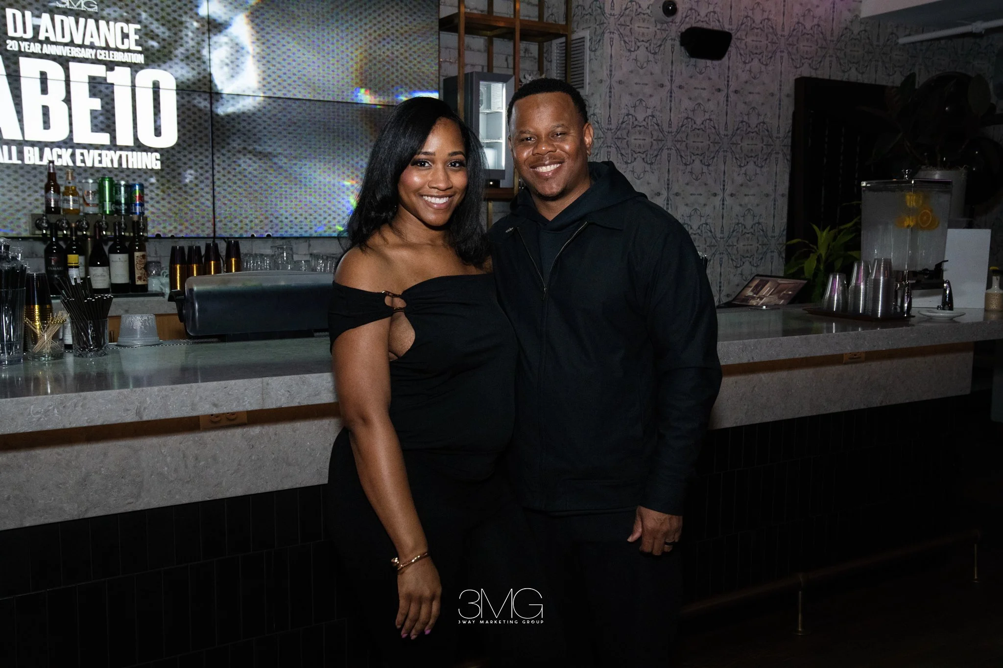 Two smiling people, a woman in a black off-shoulder dress and a man in a black jacket, stand together in front of a bar at an event or celebration. There is a large digital screen behind them with text about a DJ anniversary celebration, and various 
