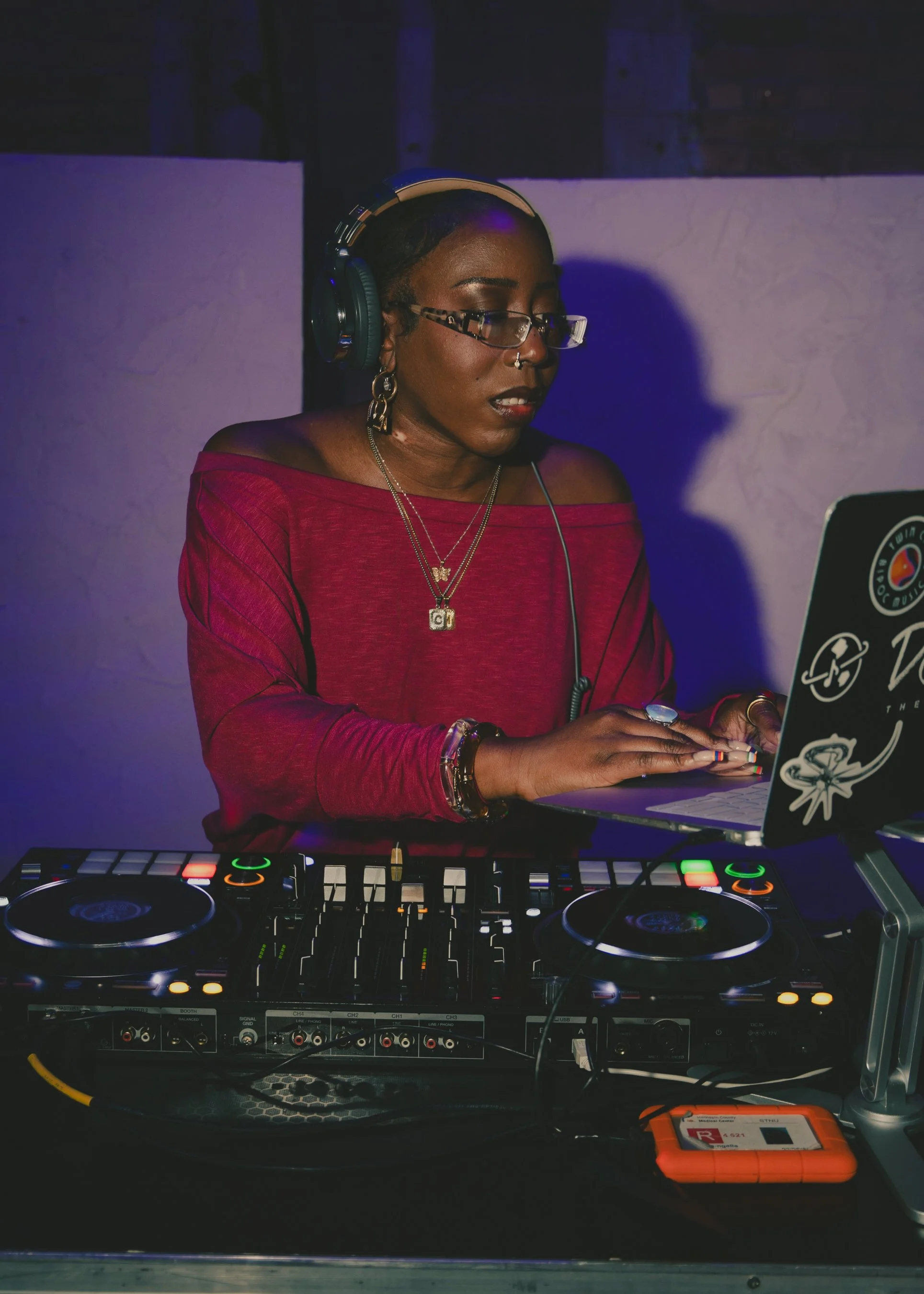 Woman DJ in a red off-the-shoulder top, wearing glasses and headphones, operating DJ equipment with mixer, turntables, and laptop decorated with stickers, in a dimly lit setting.
