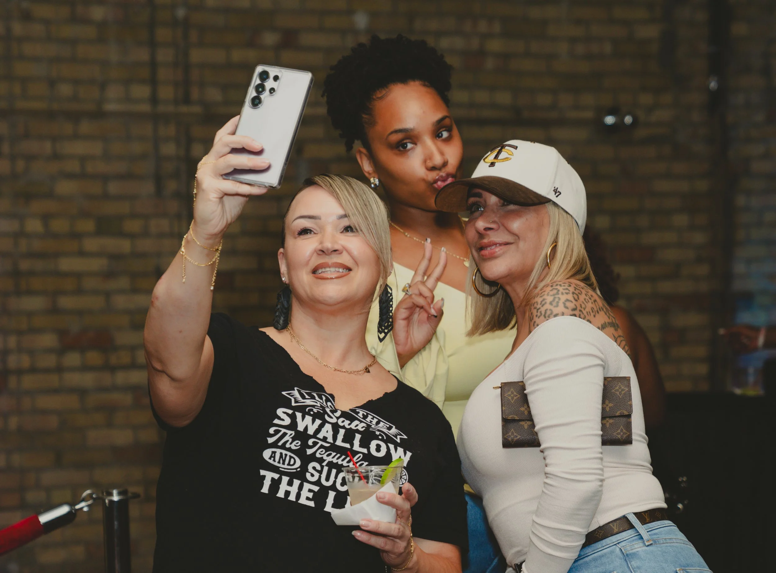 Three women taking a selfie in front of a brick wall. One woman is holding a smartphone with multiple camera lenses, and another is holding a drink. They are smiling and posing for the photo.