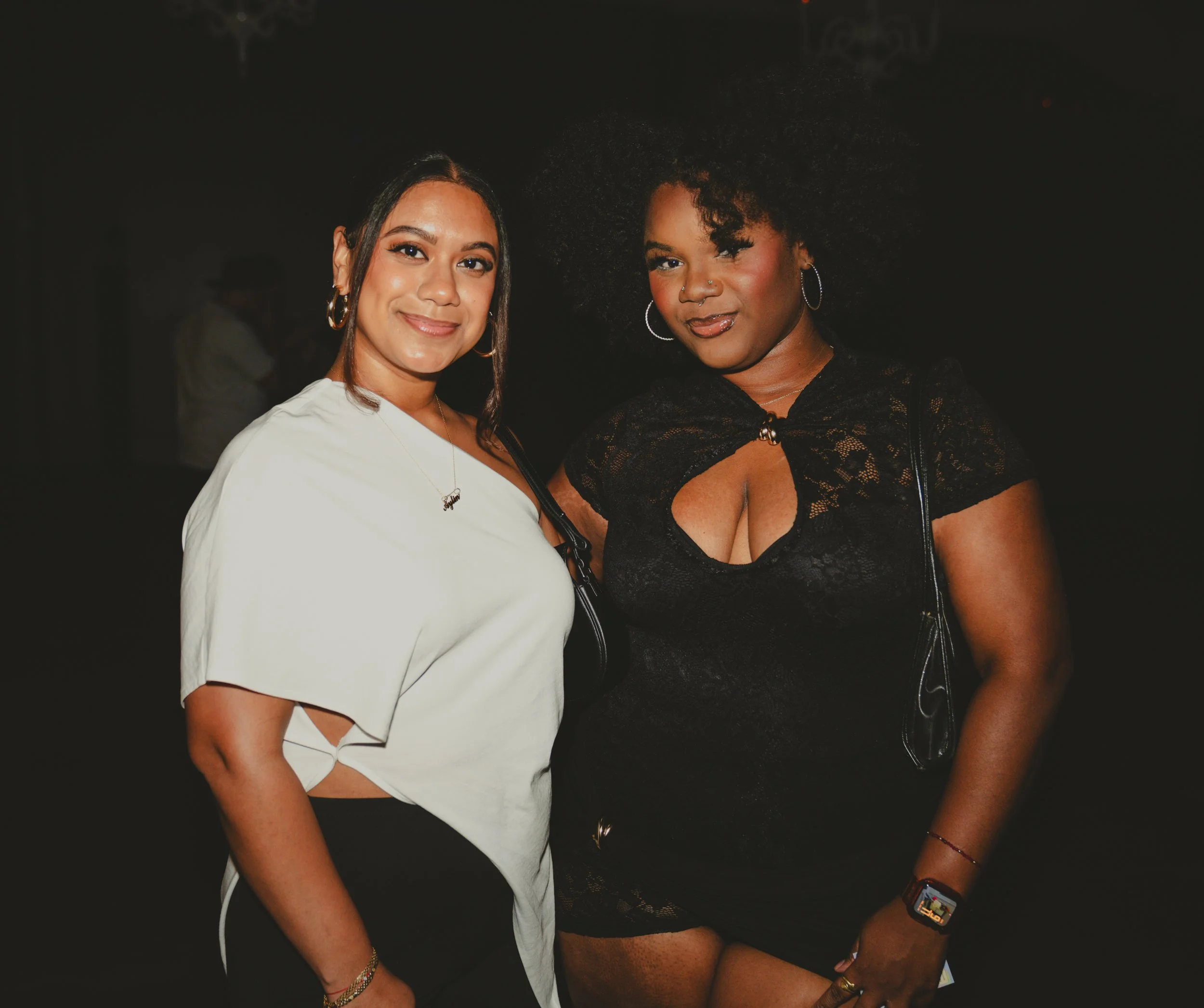 Two women standing close together, smiling at the camera. The woman on the left has long dark hair, hoop earrings, and is wearing a white top. The woman on the right has curly hair, hoop earrings, and is wearing a black lace top.