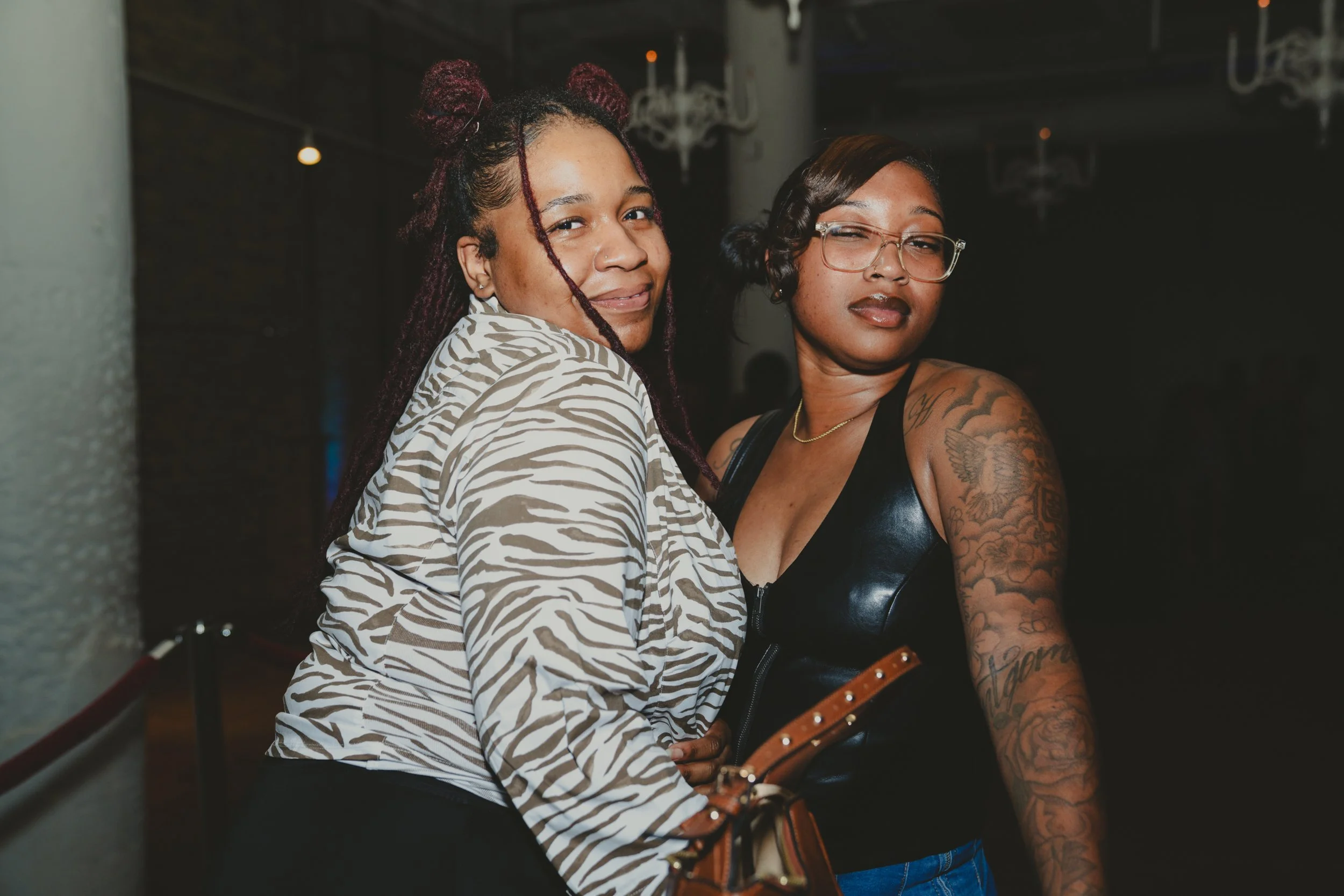 Two women with tattoos at a social event. The woman on the left has burgundy dreadlocks tied into buns and is smiling slightly, wearing a zebra-print shirt. The woman on the right has short dark hair, glasses, and tattoos on her arms, wearing a black