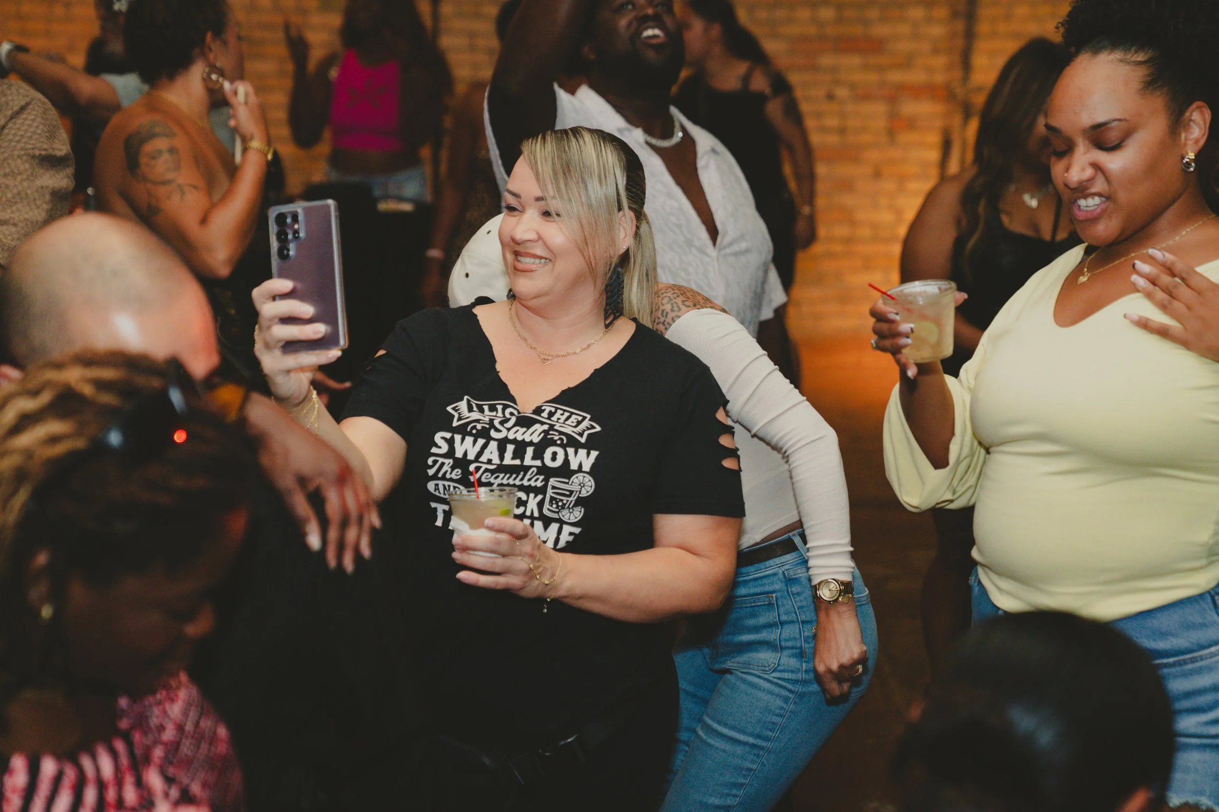 Group of diverse women dancing and enjoying drinks at a social gathering with a brick wall background.