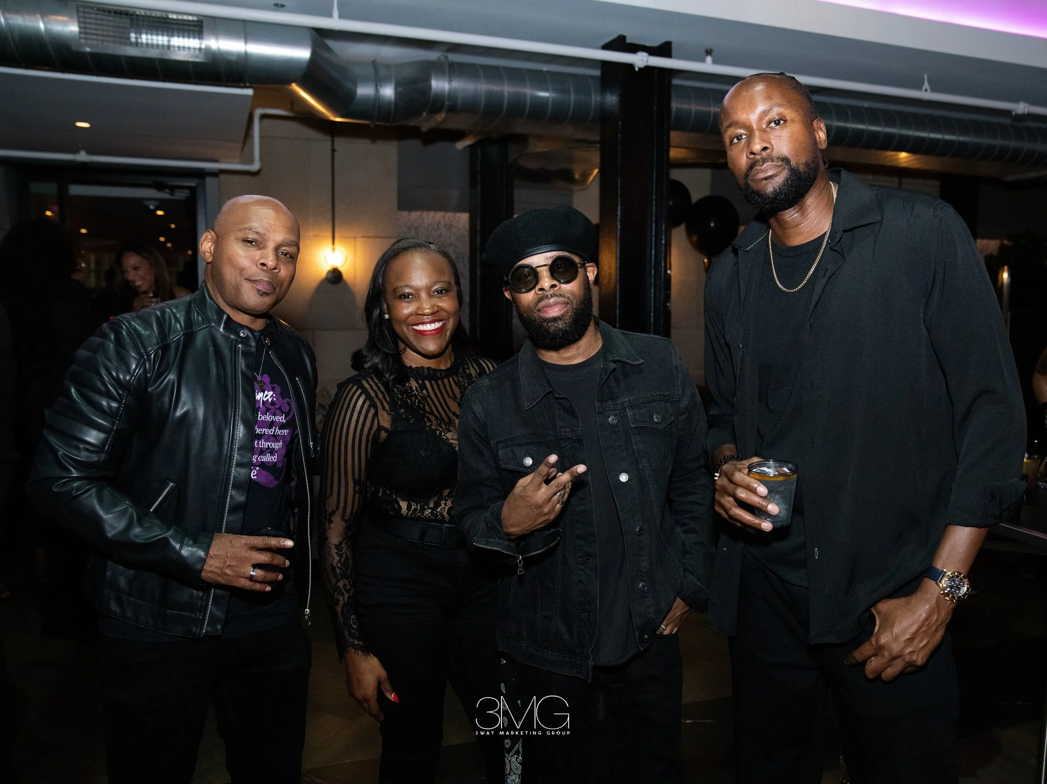 Four people standing together at an indoor event, three men and one woman, smiling and posing for the photo. The man on the far left is wearing a black leather jacket, the woman is in the middle wearing a black dress with lace details, the man next t