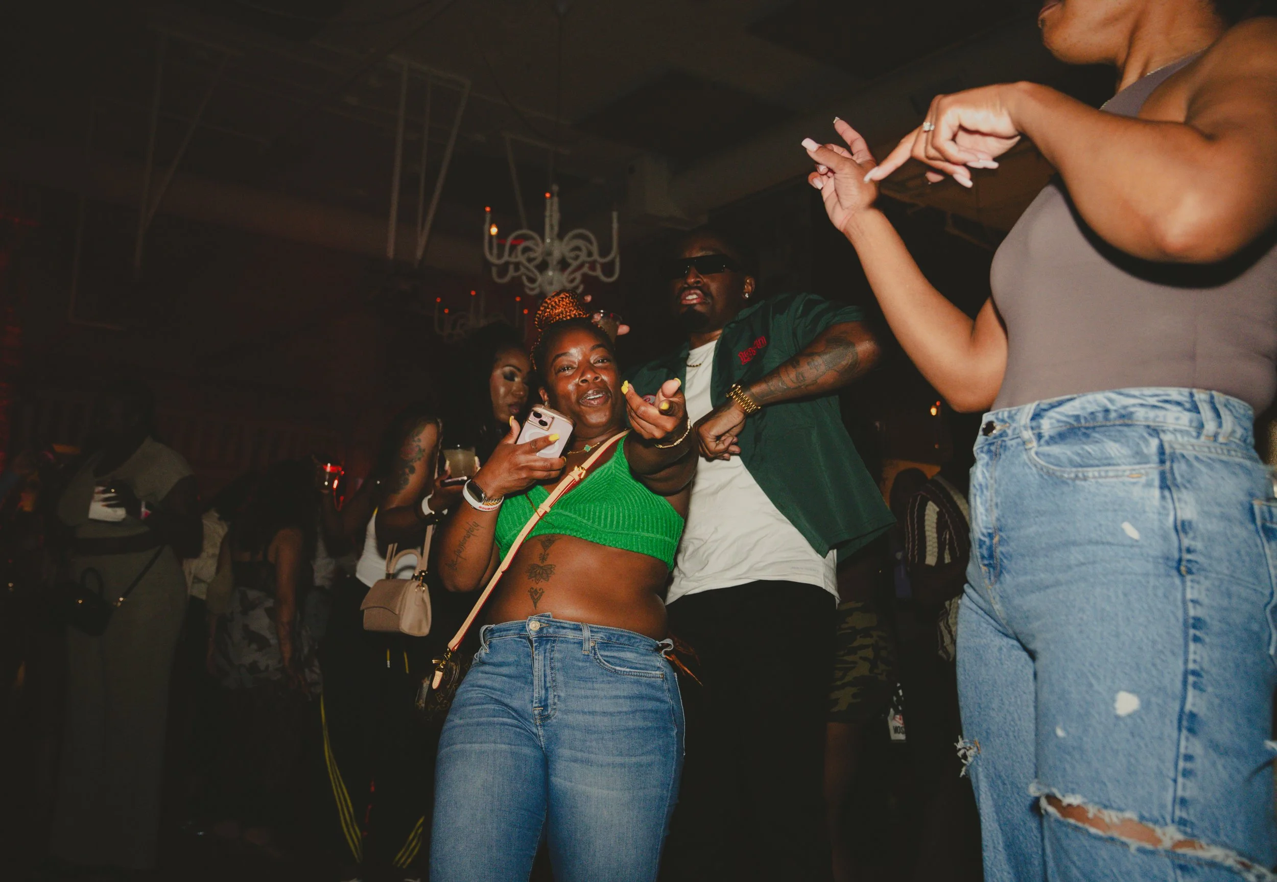 Group of people dancing at a party or nightclub, with some women holding drinks and a woman in a green top showing tattoos on her stomach.