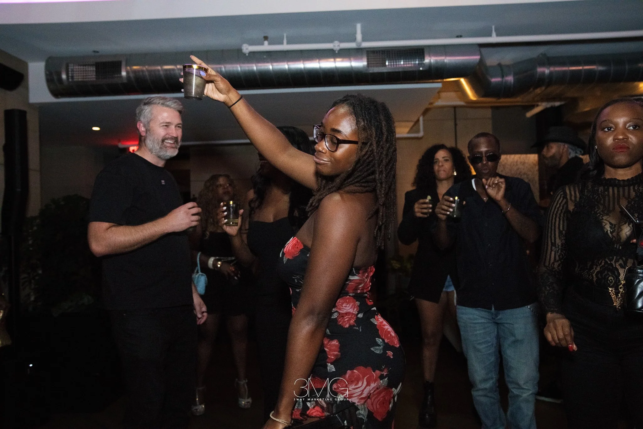 Group of people dancing and socializing in a dimly lit indoor venue, with a woman in the foreground wearing glasses and a floral dress, raising a drink.