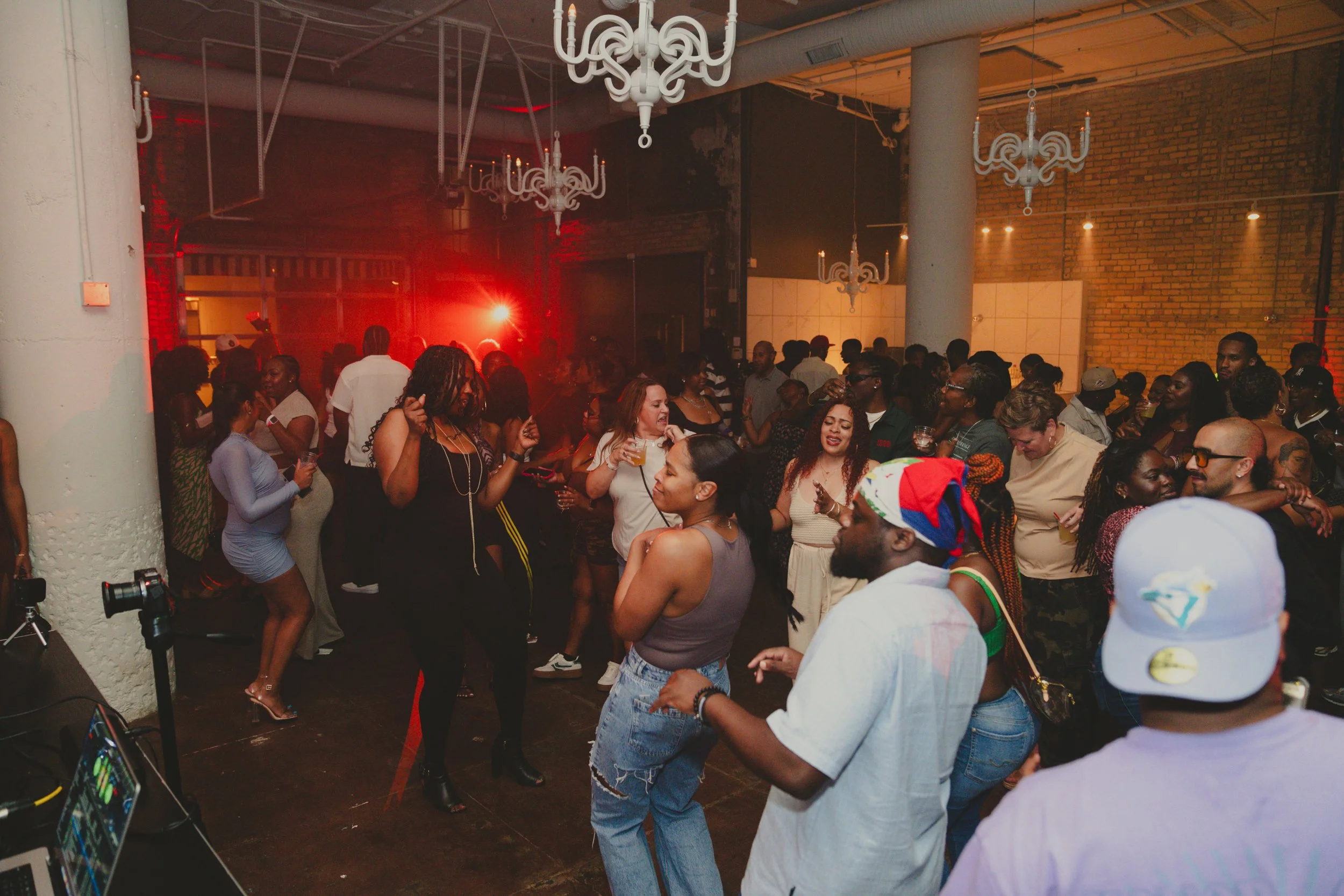 Party scene with people dancing and socializing in a dimly lit venue with exposed brick walls and chandeliers, red lights, and a DJ setup.