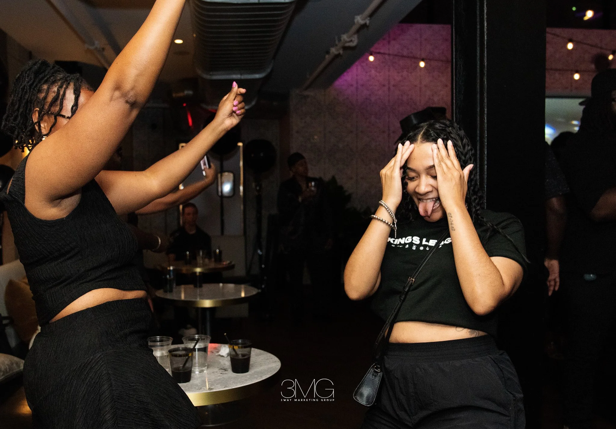 Two young women with braided hair and wearing black tops are at a social gathering or party, with one smiling and holding her head with her hands, while the other is raising her arm, possibly dancing. There are drinks on a table and other people in t
