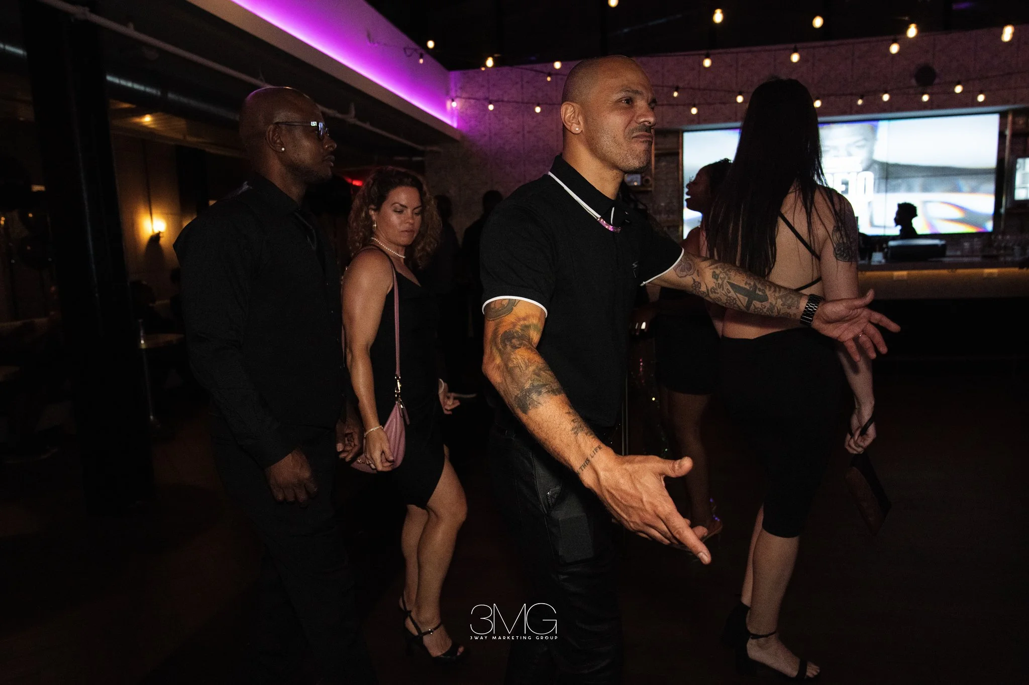 A group of casually dressed people dancing and socializing in a dimly lit bar or nightclub with purple and yellow lighting. A man with tattoos is in the foreground, and a large screen displaying colorful visuals is in the background.