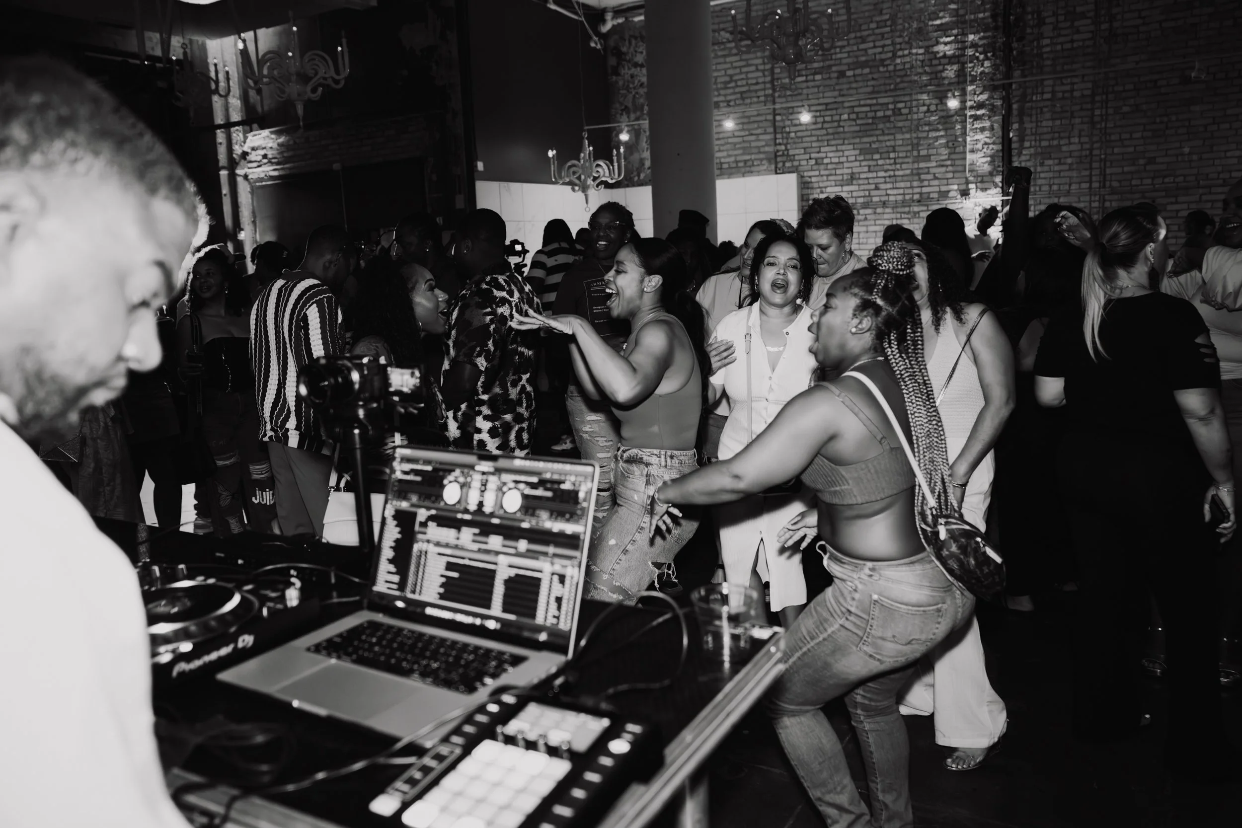 People dancing and socializing at a party or event in a venue with brick walls and chandeliers.