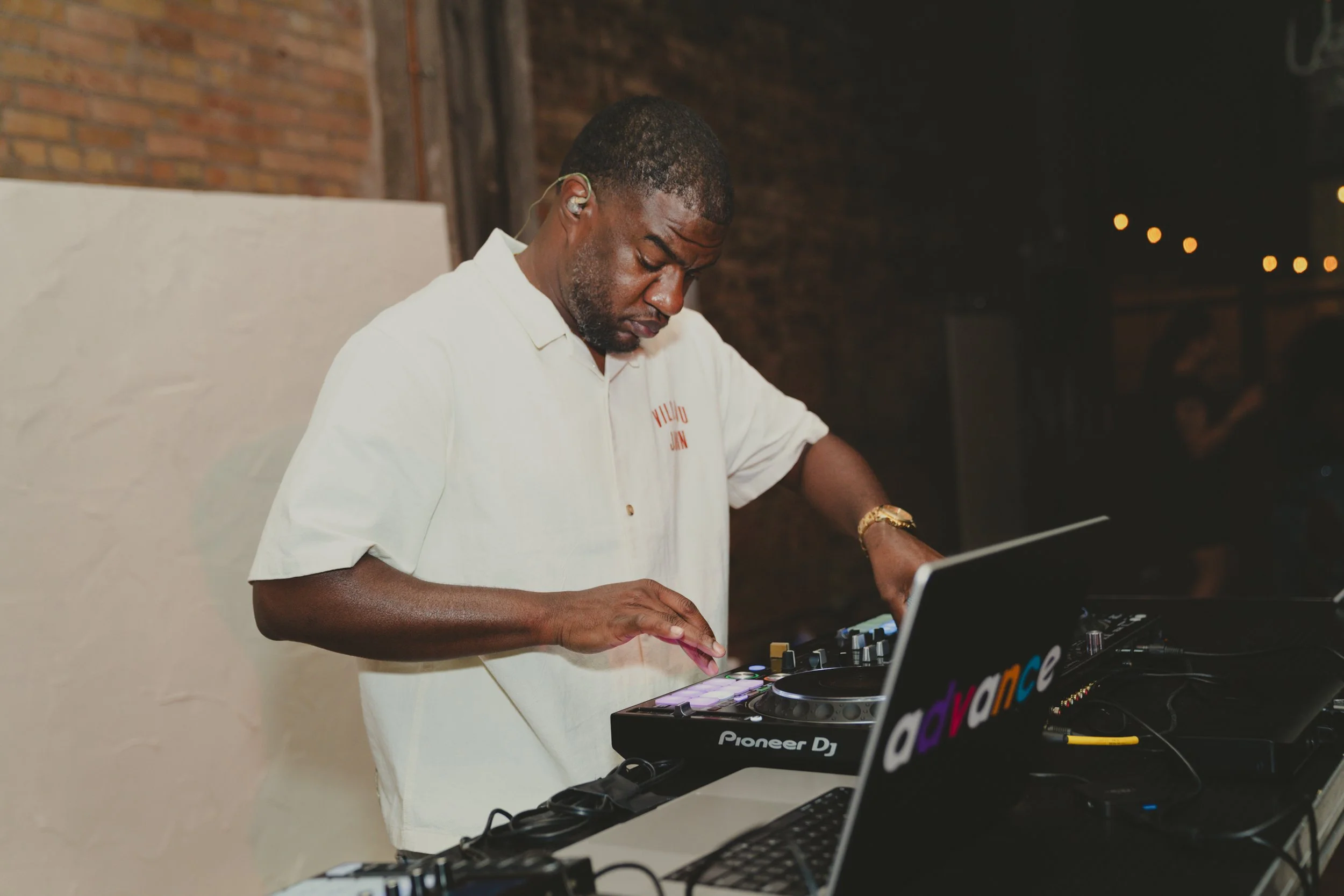 An African American man wearing a white shirt, gold watch, and in-ear headphones, DJing with a Pioneer DJ controller in a dimly lit indoor setting with a brick wall background.