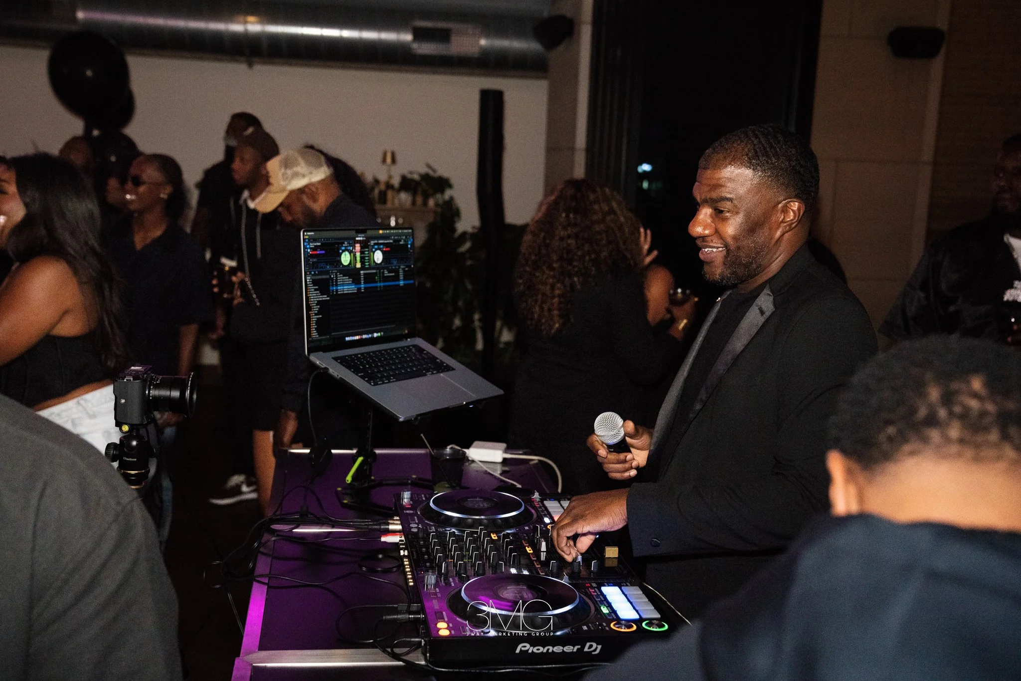 A DJ in a black suit with a microphone, operating DJ equipment at a nightclub with people dancing and socializing in the background.