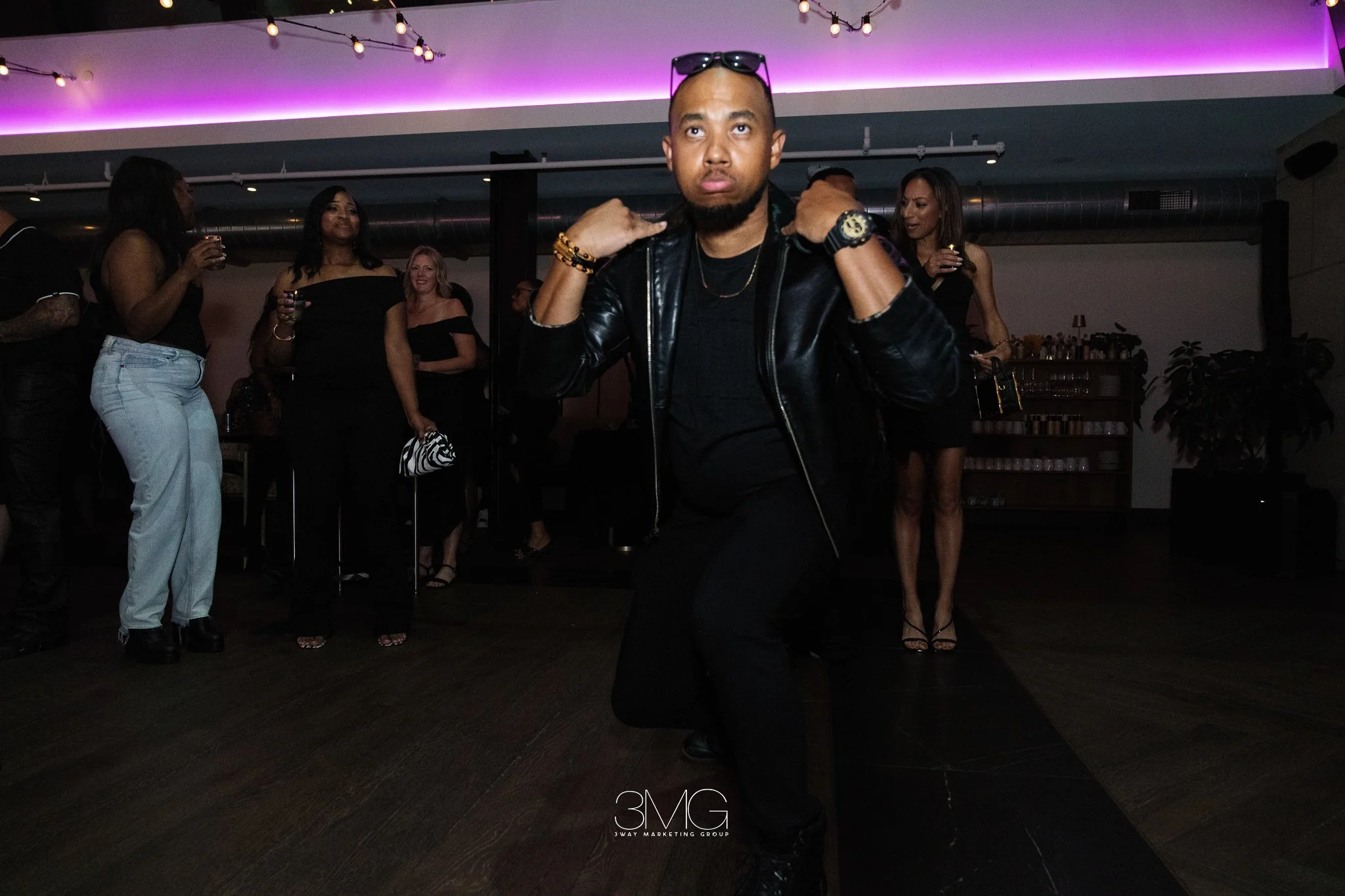 A group of people at a party, with a man in the foreground wearing a leather jacket, sunglasses on his head, and accessories, confidently posing. The background shows women in black outfits holding drinks, enjoying the event, with purple lighting and