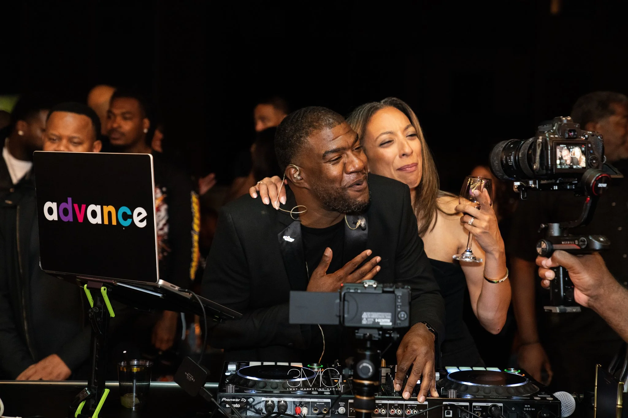 A man and a woman are standing close together at a party, smiling at the camera. The man is wearing a black suit, and the woman is holding a glass of wine. They are surrounded by people, and there are DJ equipment and a laptop with the logo 'advance'