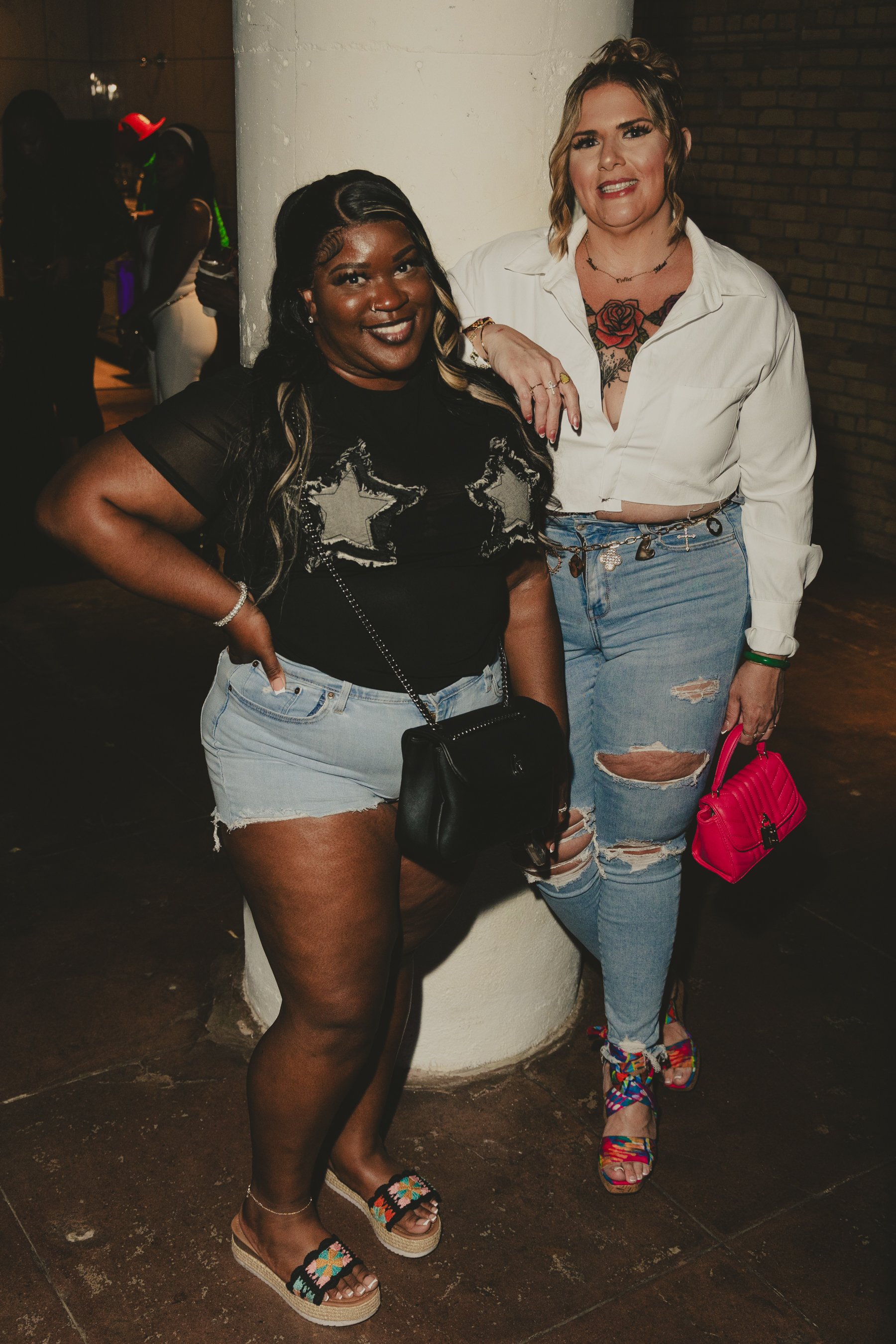 Two women standing next to each other, one wearing a black top with star patterns and light denim shorts, and the other wearing a white shirt with ripped jeans and colorful sandals. They are indoors, with other people in the background.