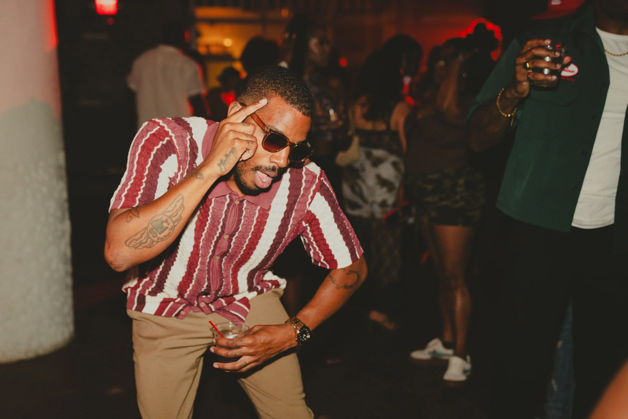 A young man with tattoos, wearing sunglasses, a patterned shirt, and beige pants, is dancing at a party while holding a drink with a red straw. There are people in the background dancing and socializing in a dimly lit venue.