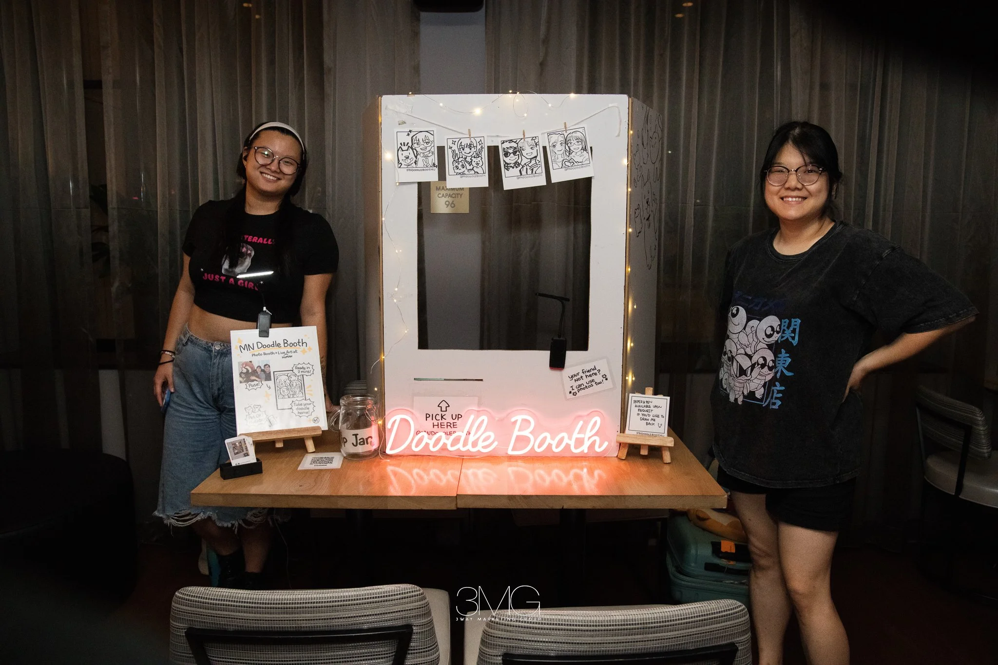 Two women standing on either side of a photo booth with a sign that says "Doodle Booth" at a night event. The woman on the left is smiling, wearing glasses and a black crop top with jeans. The woman on the right is also smiling, wearing glasses and a