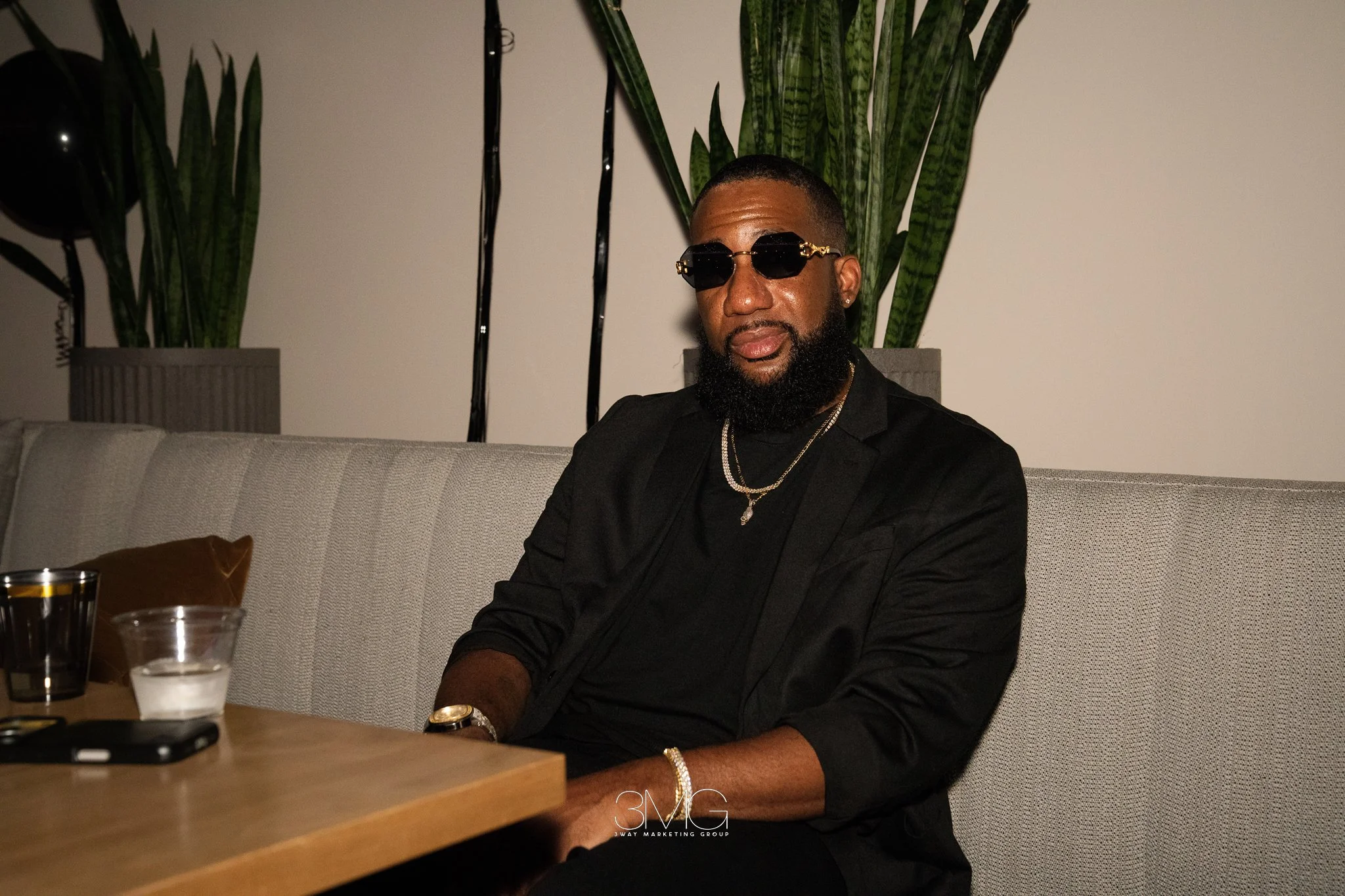 A man with a beard and short hair wearing stylish black sunglasses, a black shirt, gold chains, and a watch sitting on a beige couch in a room with large green plants in the background.