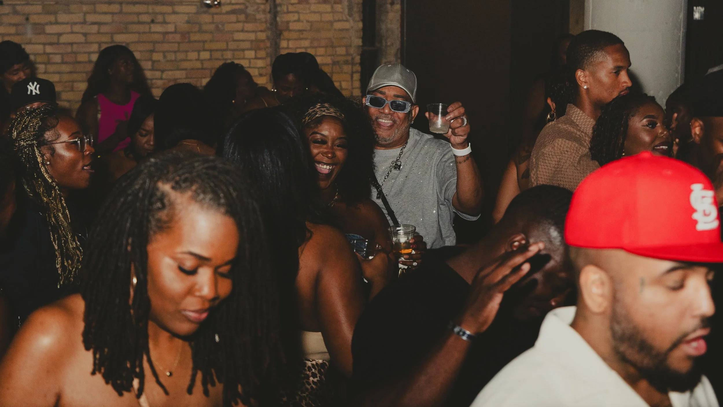 A diverse group of people enjoying a social gathering in a dimly lit indoor space with brick walls, some holding drinks, smiling and engaging with each other.