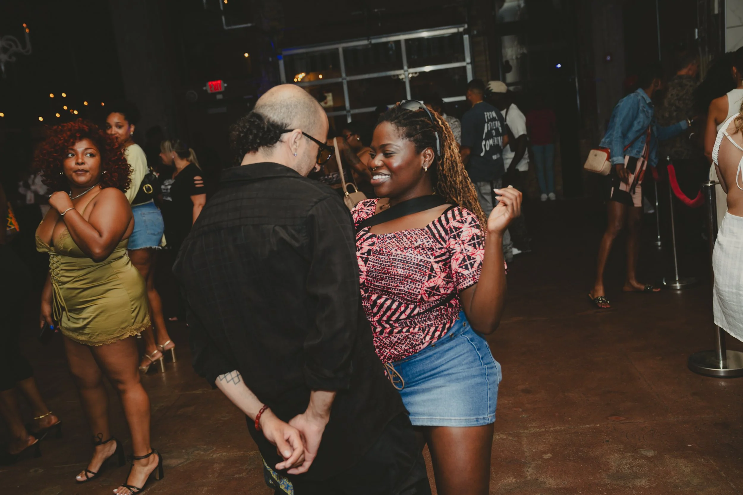 People dancing and socializing in a dark indoor venue with dim lighting, some holding hands and others standing in groups.