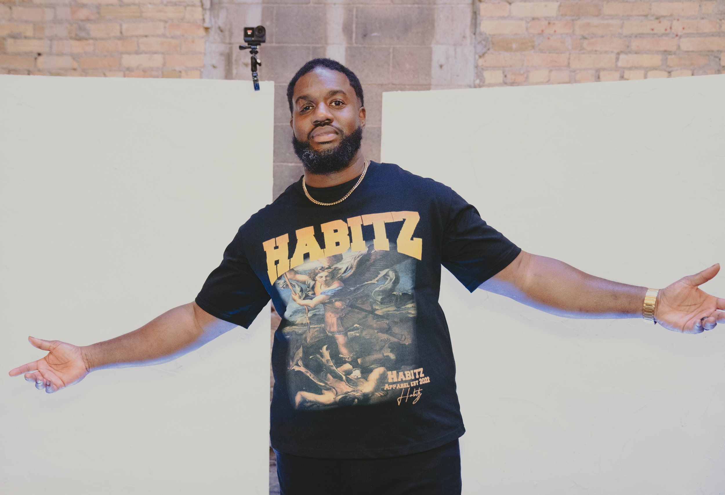 A man with a beard wearing a black graphic t-shirt and a gold chain, standing with arms outstretched against a partially obscured background with a brick wall and white paper or boards.