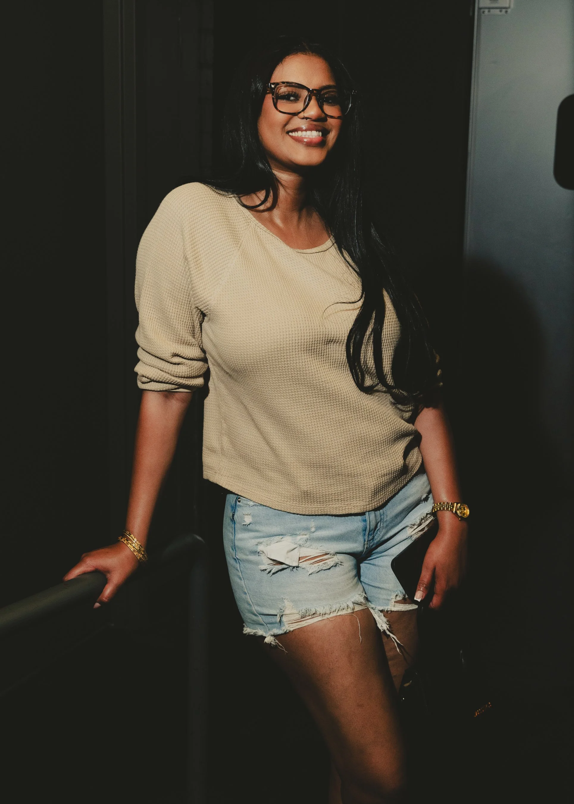 A woman with long black hair, glasses, and a nose piercing, smiling, wearing a beige sweater and ripped denim shorts, holding onto a railing, standing in a dark setting.