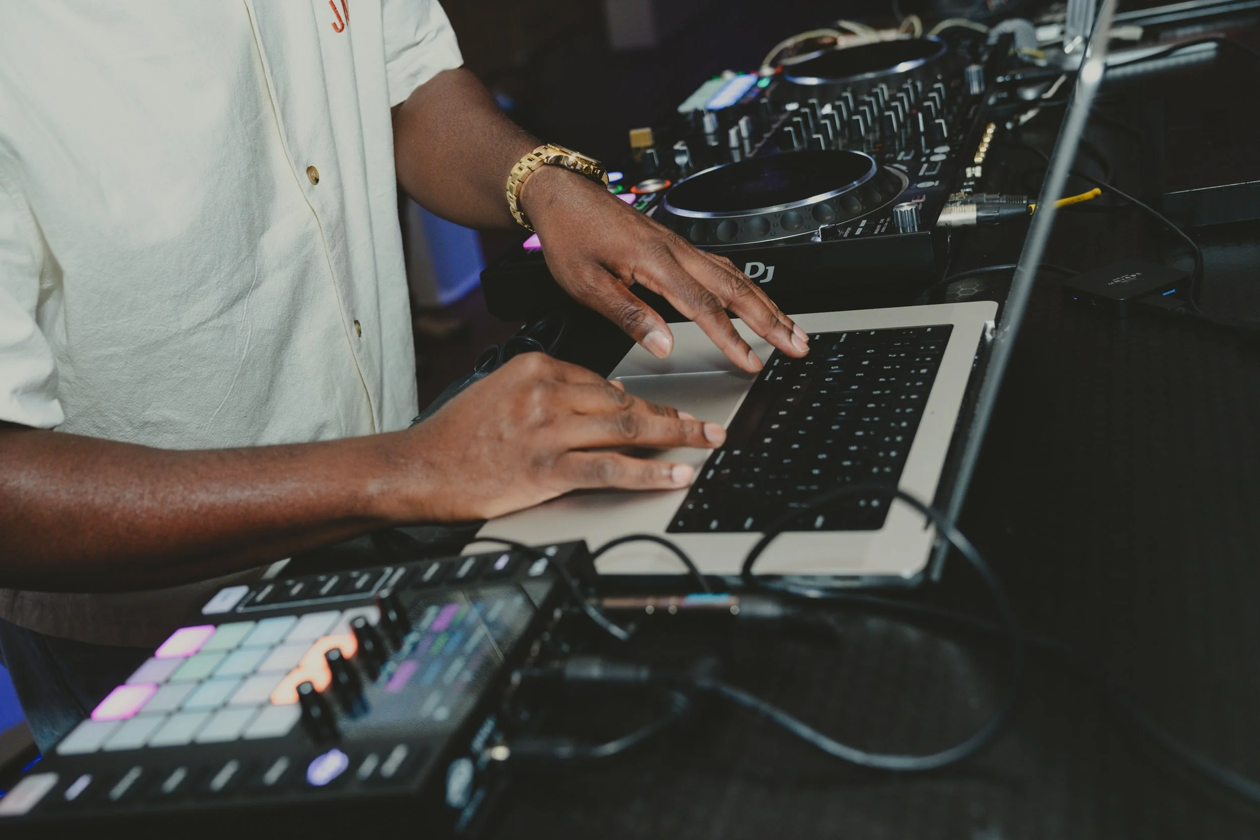 A DJ wearing a white shirt and gold bracelet operating DJ equipment with a laptop, mixer, and colorful pad controller.