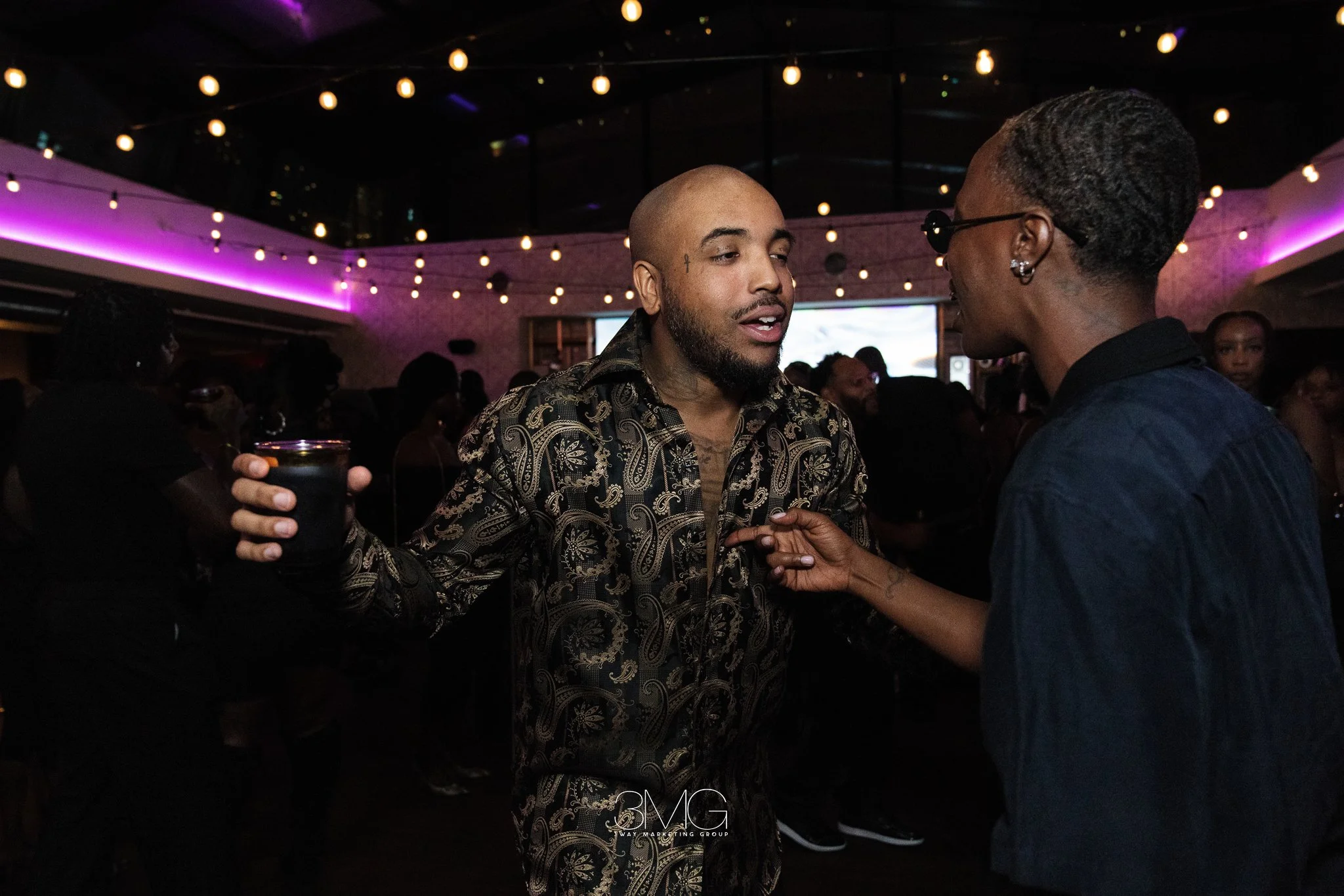 Two men having a conversation at a party with string lights and purple lighting in the background.